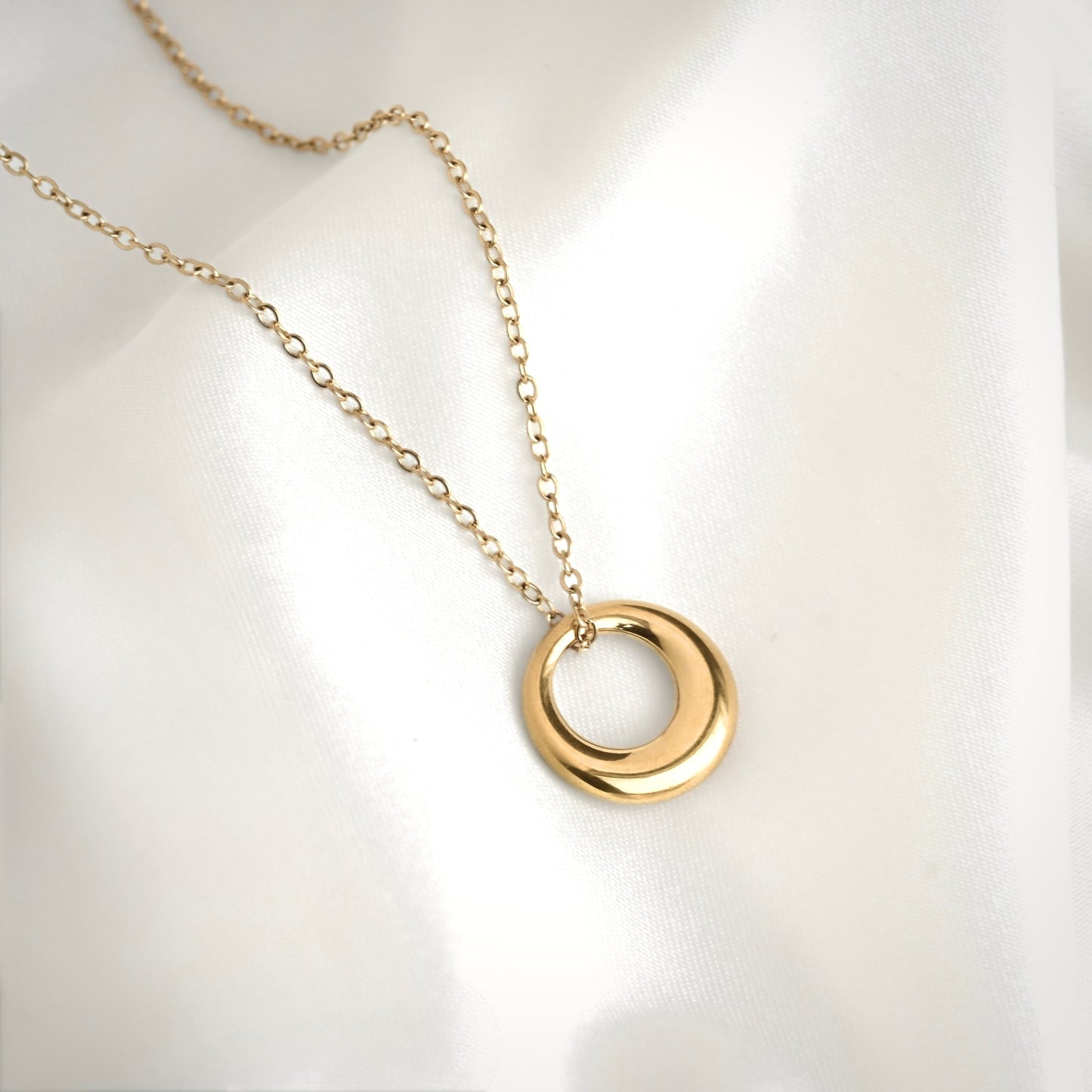 A gold chain necklace with a polished, open circular pendant, displayed on a soft, white satin fabric background.