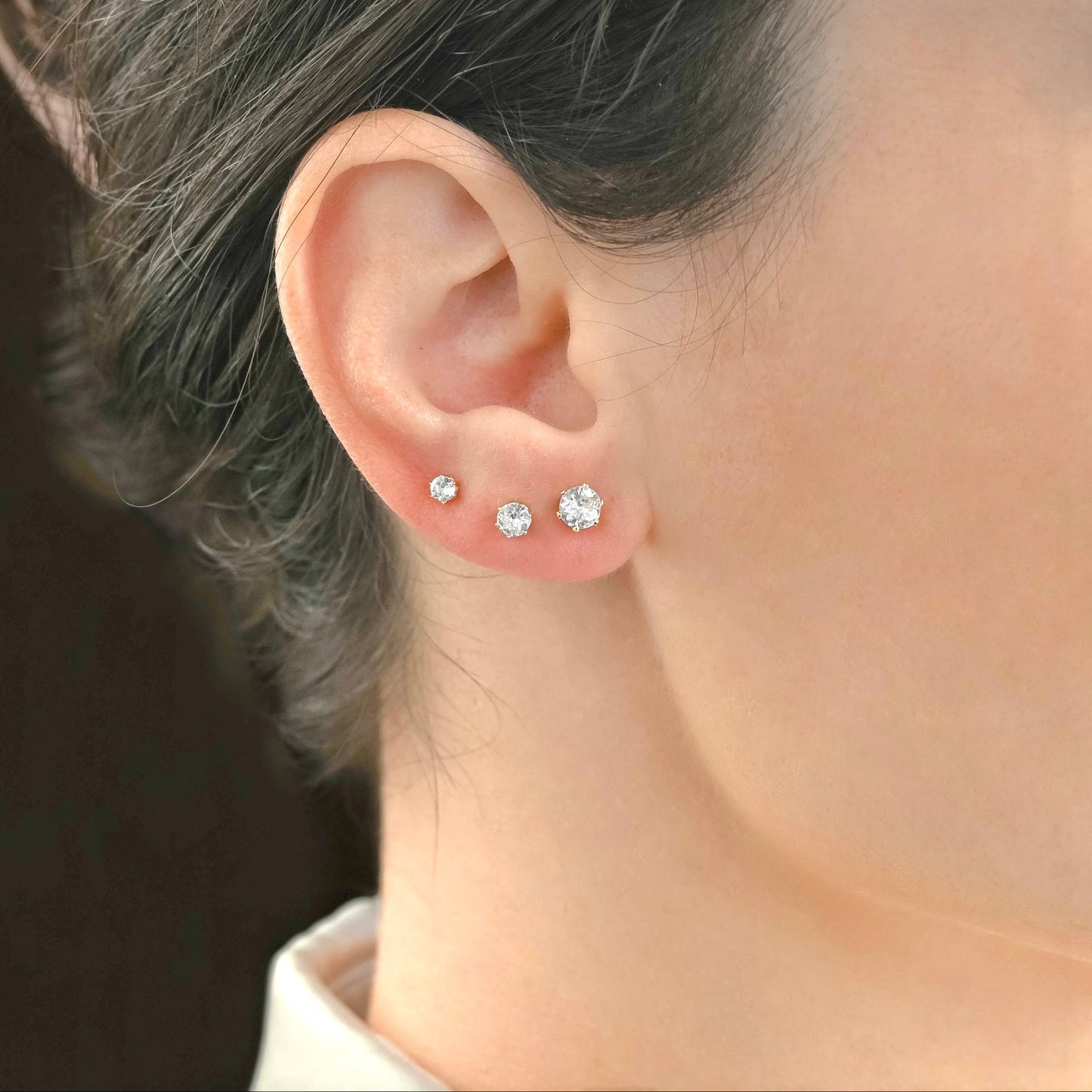 Close-up of an ear wearing three round diamond stud earrings in 3mm, 4mm and 5mm with a blurred background