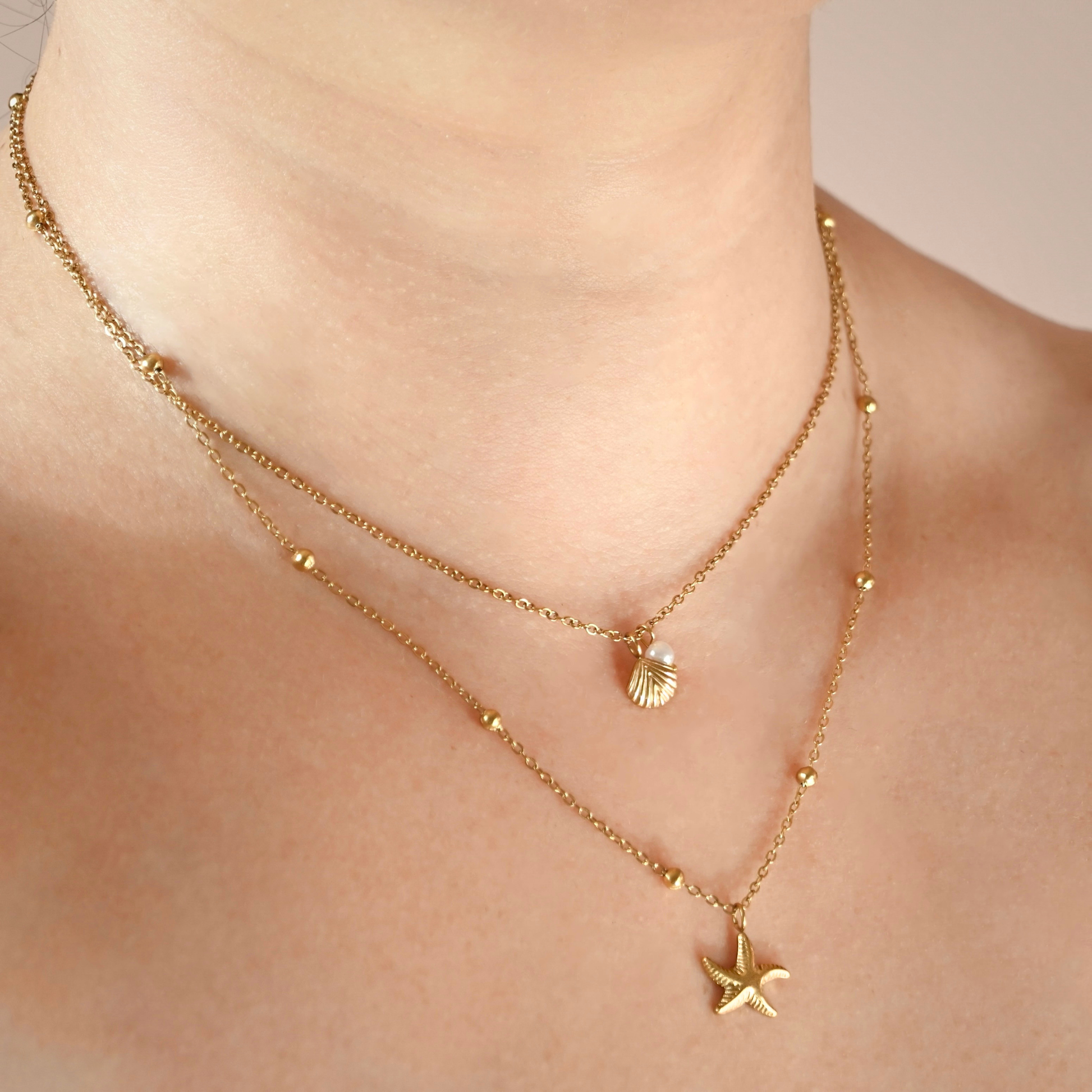 A close-up of a model's neck and upper chest wearing a delicate layered gold necklace with a small shell pendant and a sea star pendant.
