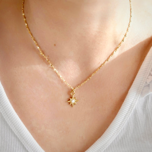 A close-up of a person wearing a delicate gold chain necklace with a small gold starburst pendant featuring a clear central stone, styled with a white ribbed V-neck top.