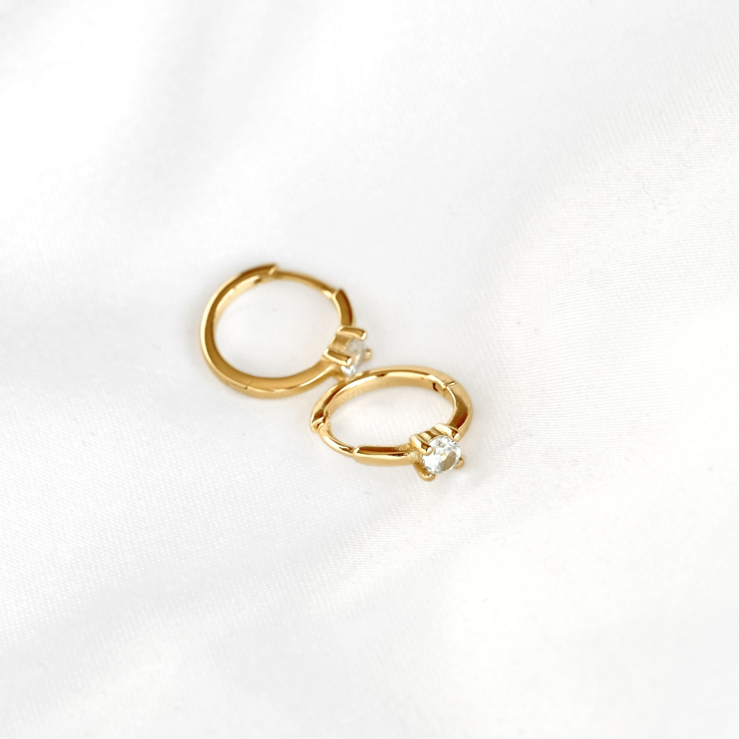 A pair of small gold hoop earrings with a single clear round gemstone in each earring, displayed on a soft white fabric background.