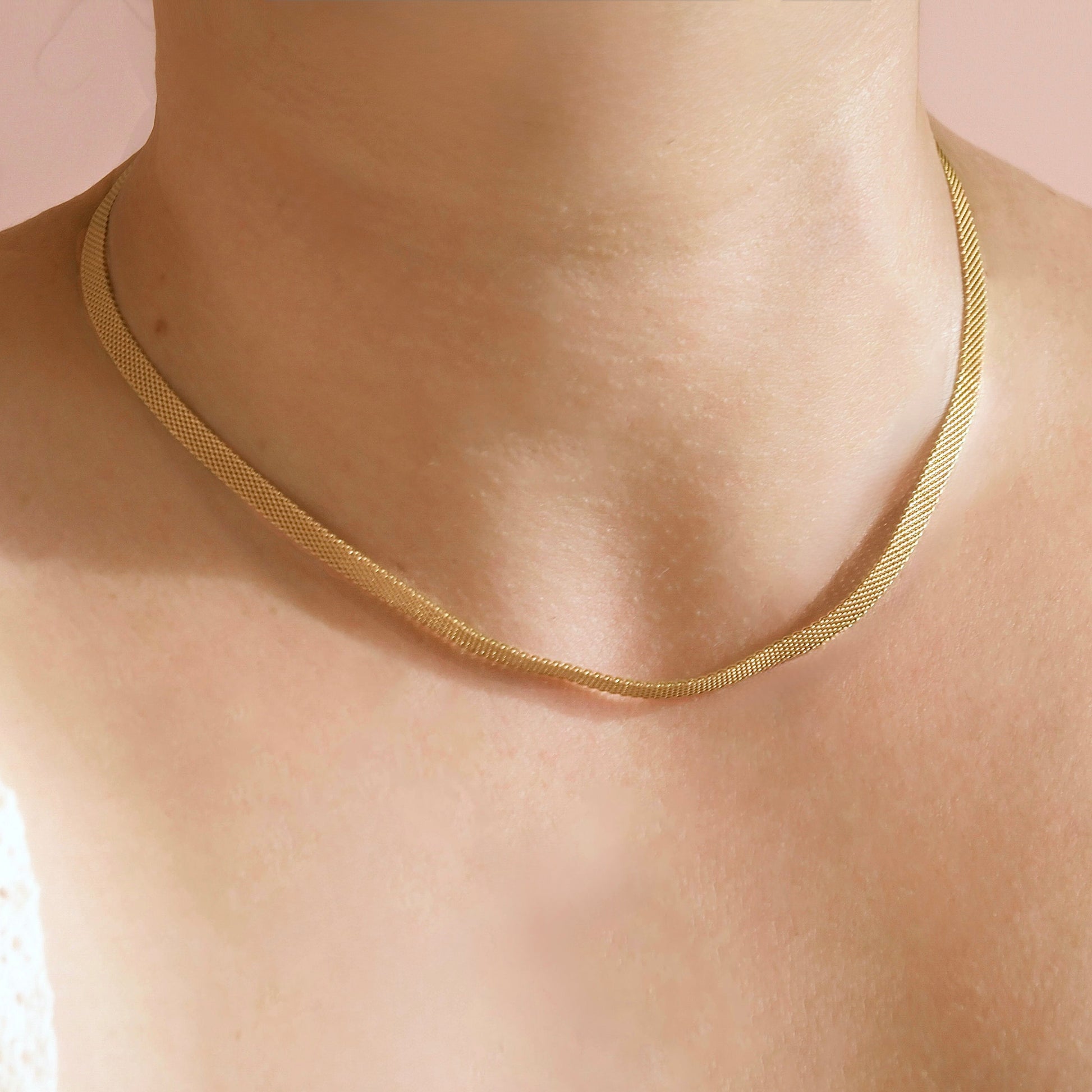 A close-up of a person's neck and upper chest wearing a delicate gold snake chain necklace against a soft pink background.