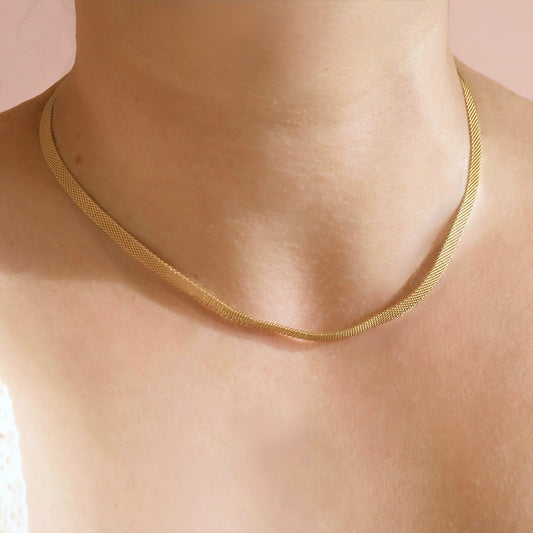 A close-up of a person's neck and upper chest wearing a delicate gold snake chain necklace against a soft pink background.