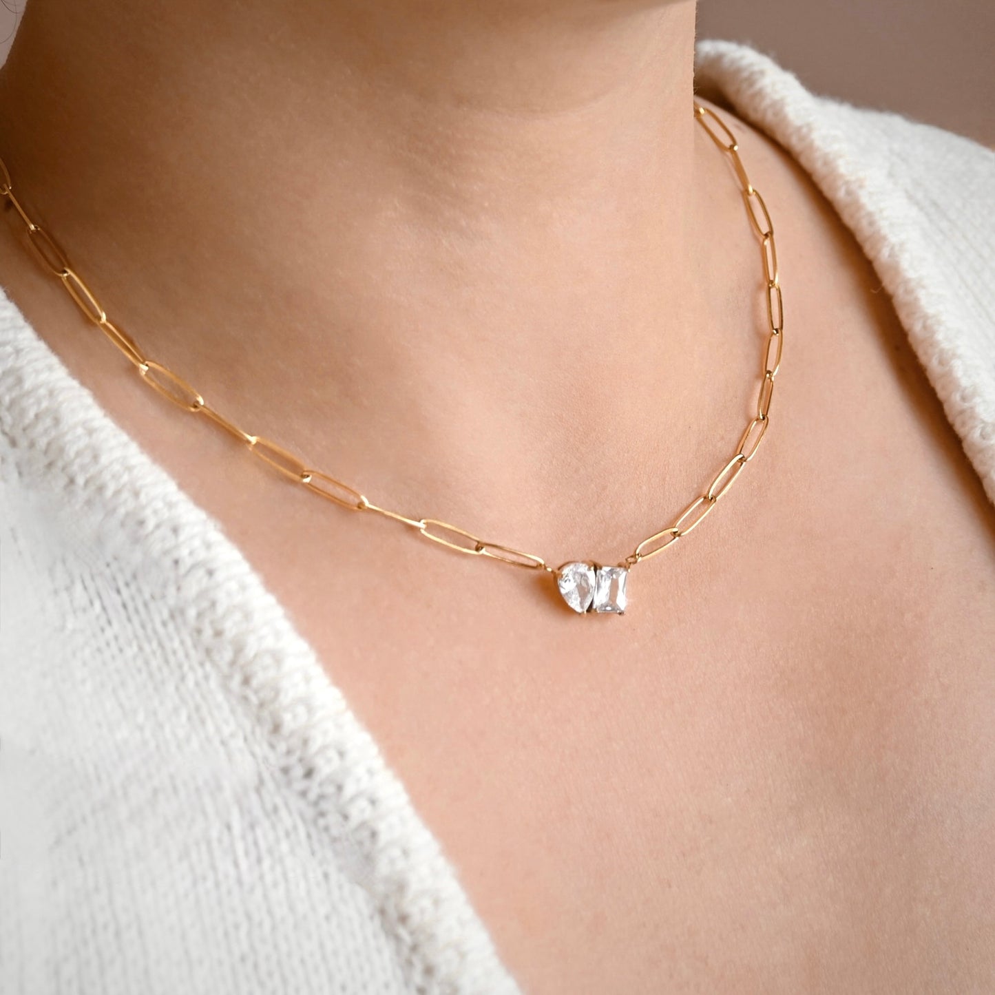 A close-up of a model wearing a delicate gold  paperclip chain necklace with a small cluster of clear gemstones at the center, styled with a white knit top.
