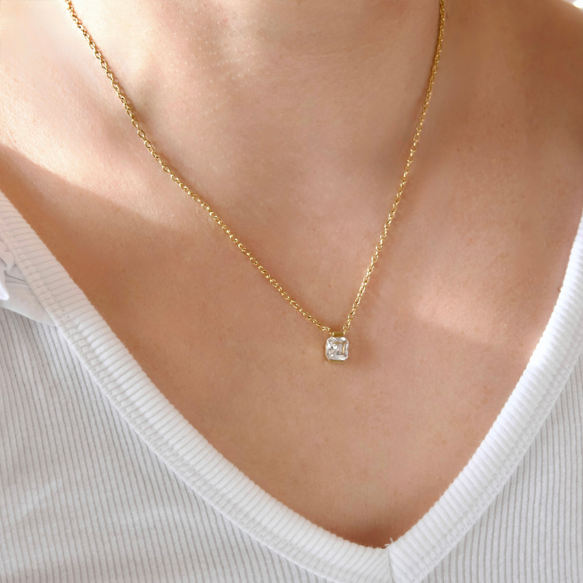 A delicate gold chain necklace with a small square clear gemstone pendant, worn on a model's neck over a white ribbed V-neck top.