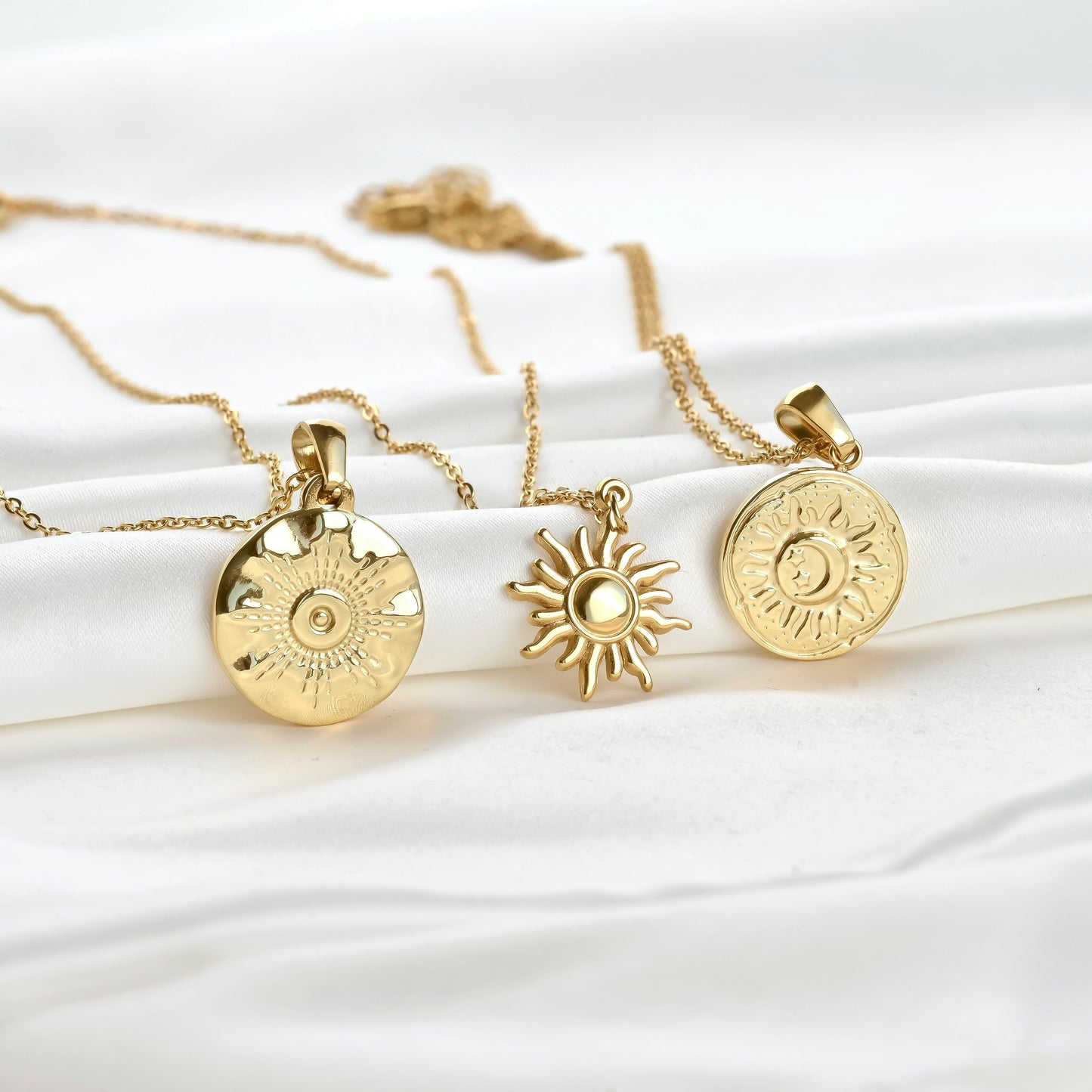 Three gold necklaces with sun-shaped pendants on a white fabric background