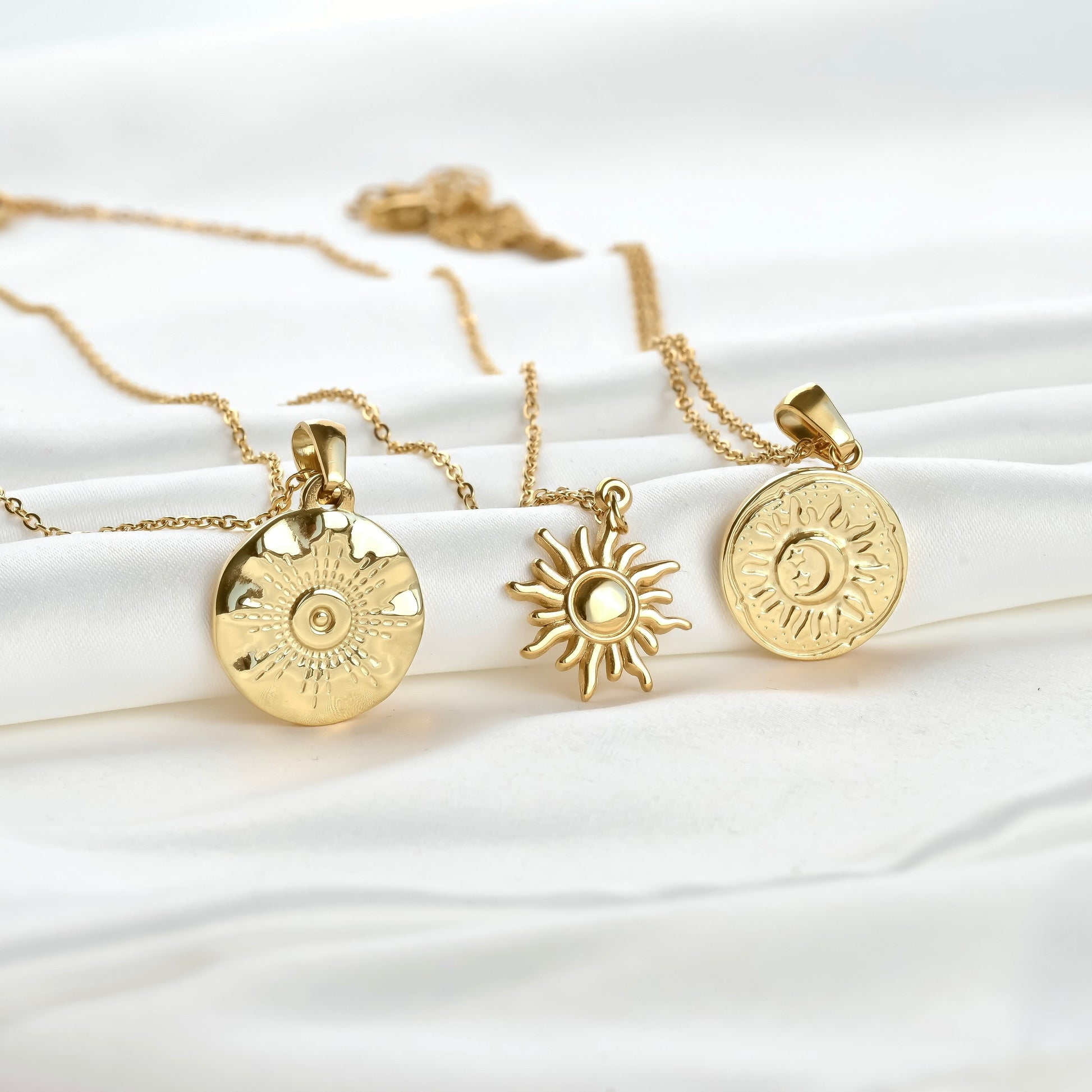 Three gold necklaces with sun-shaped pendants on a white fabric background
