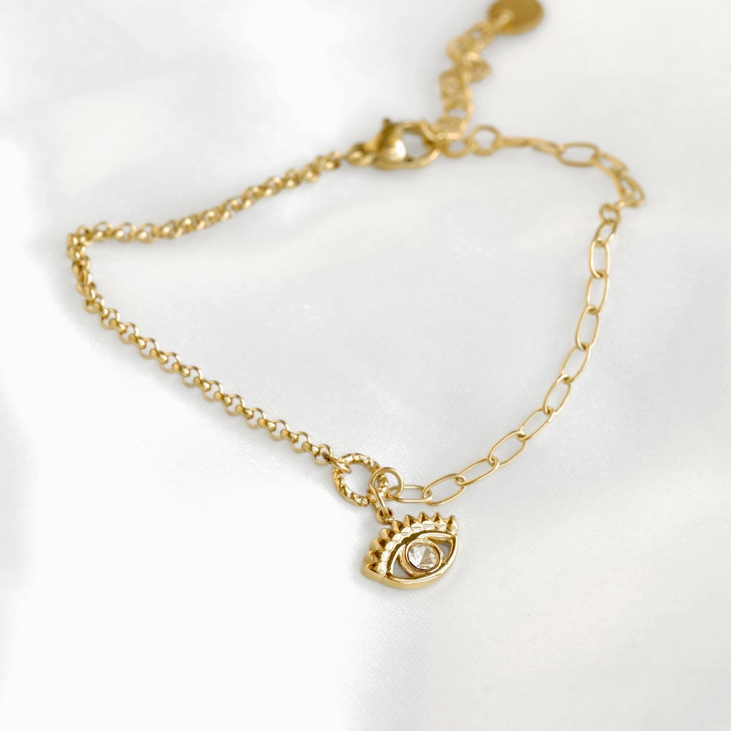 A delicate gold chain bracelet featuring a small evil eye charm with a clear central gemstone, displayed on a soft white fabric background.