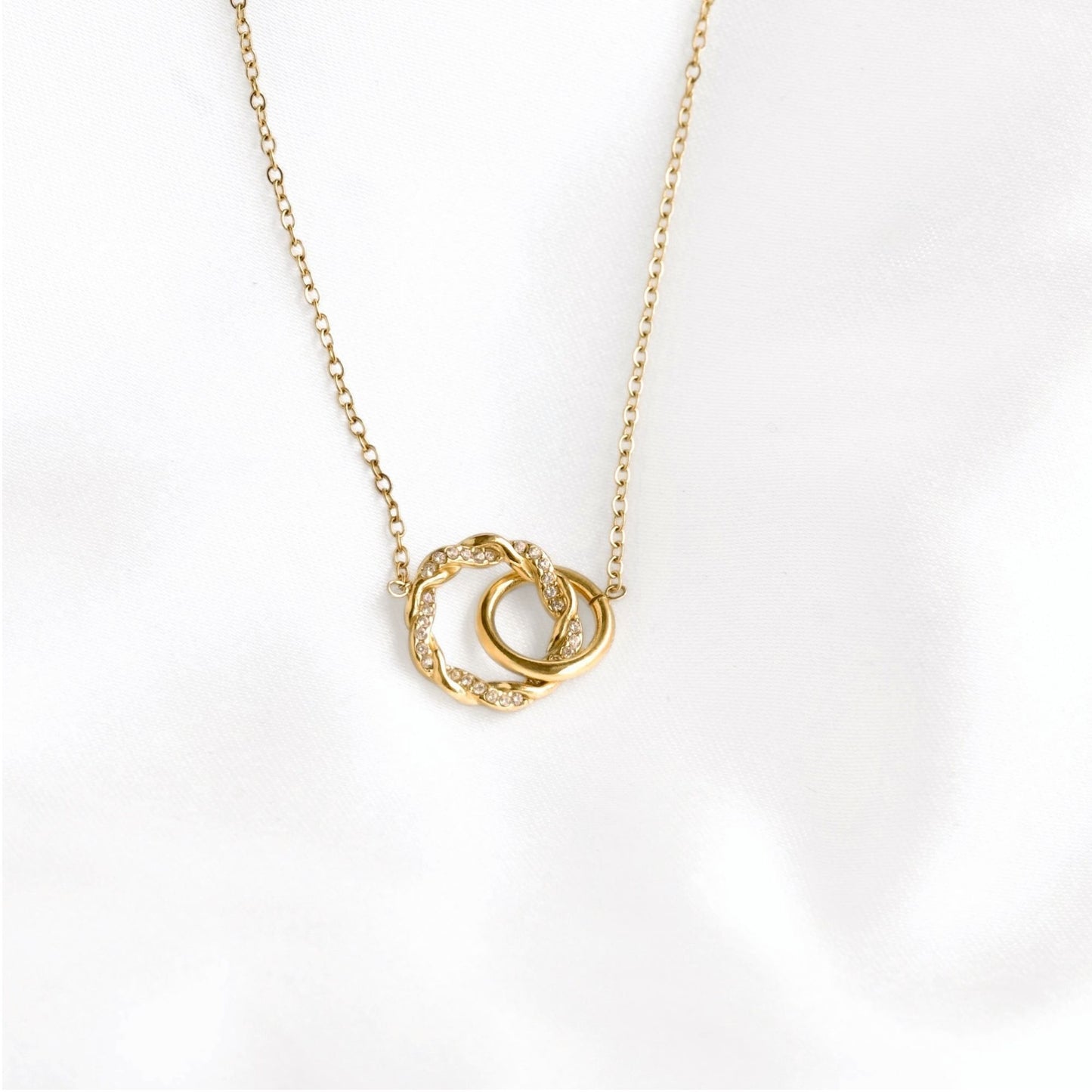 A delicate gold chain necklace featuring two interlocking circular pendant rings, one textured and one smooth, set against a soft white background.