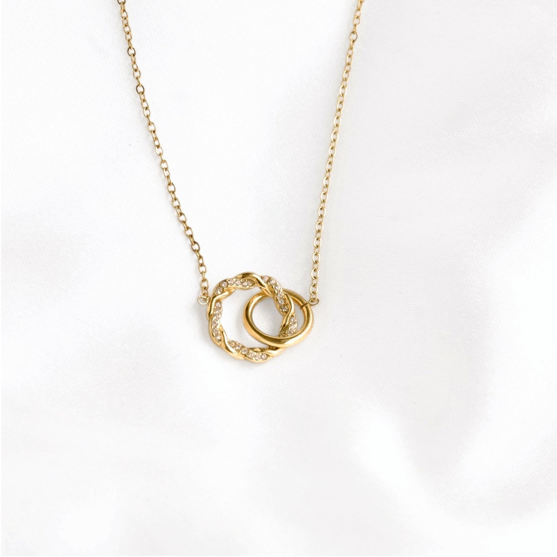 A delicate gold chain necklace featuring two interlocking circular pendant rings, one textured and one smooth, set against a soft white background.