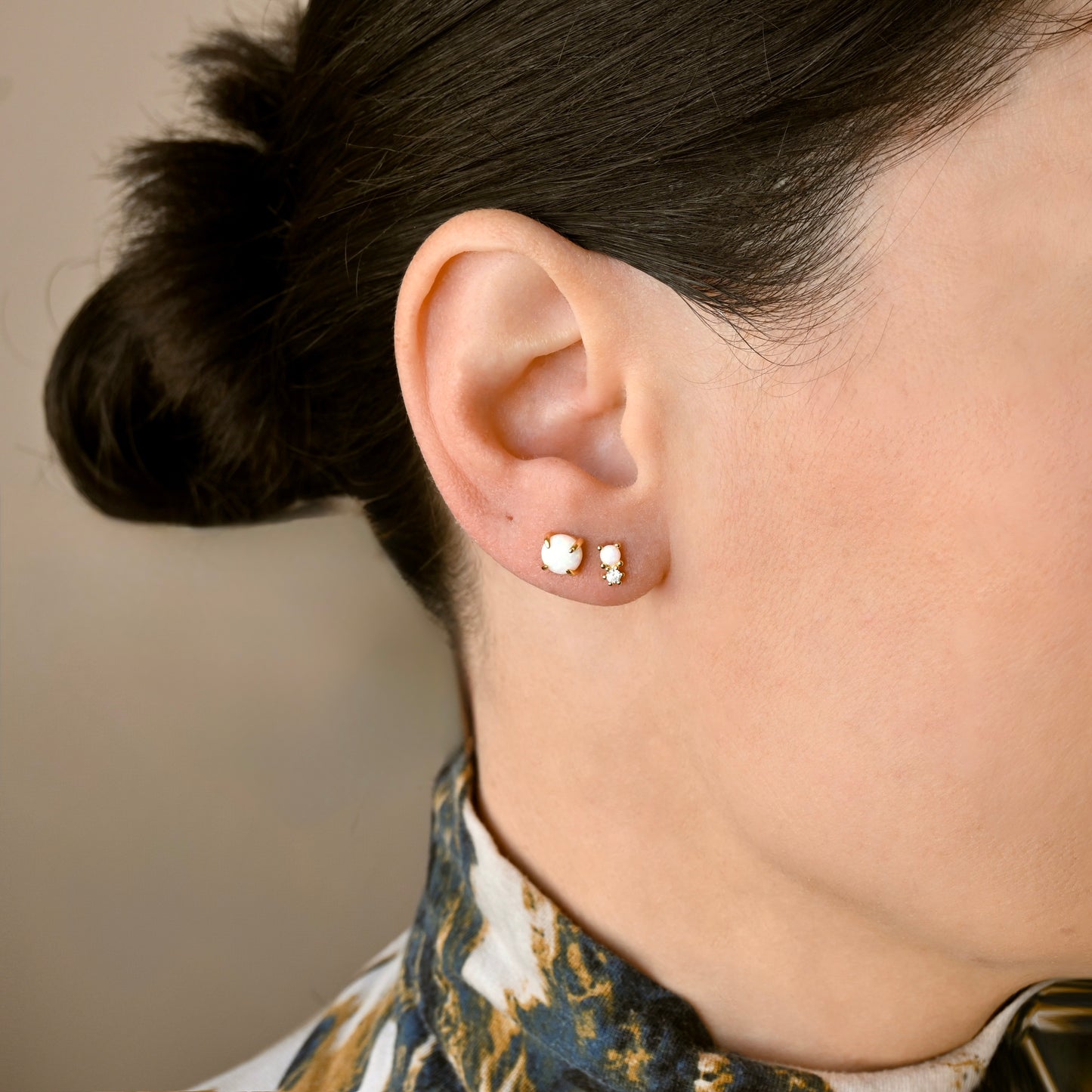 A close-up side profile of a person wearing two small gold stud earrings , one with medium opal stone and one with small opal and cz stacked on their ear, with dark hair tied back and a patterned collar visible.