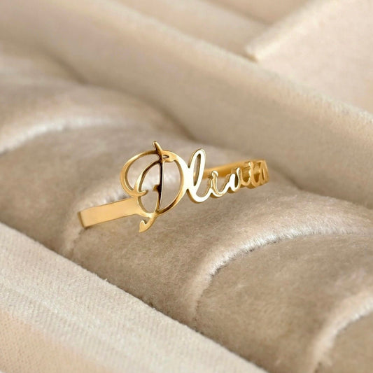 A gold name ring with the name 'Olivia' in cursive script, displayed on a beige velvet jewelry box.