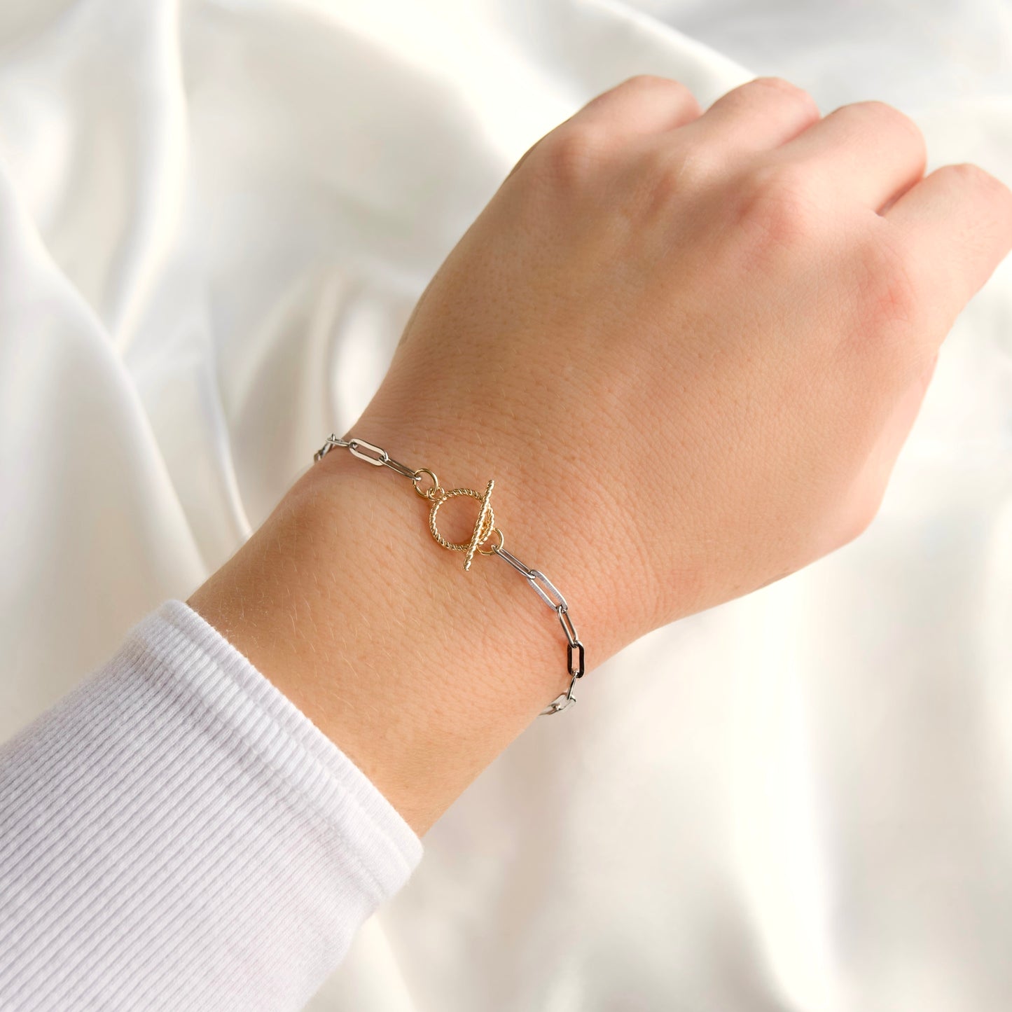 A close-up of a person's wrist wearing a delicate two-tone paperclip chain bracelet with a 10k plated twisted gold toggle clasp, set against a soft white fabric background.