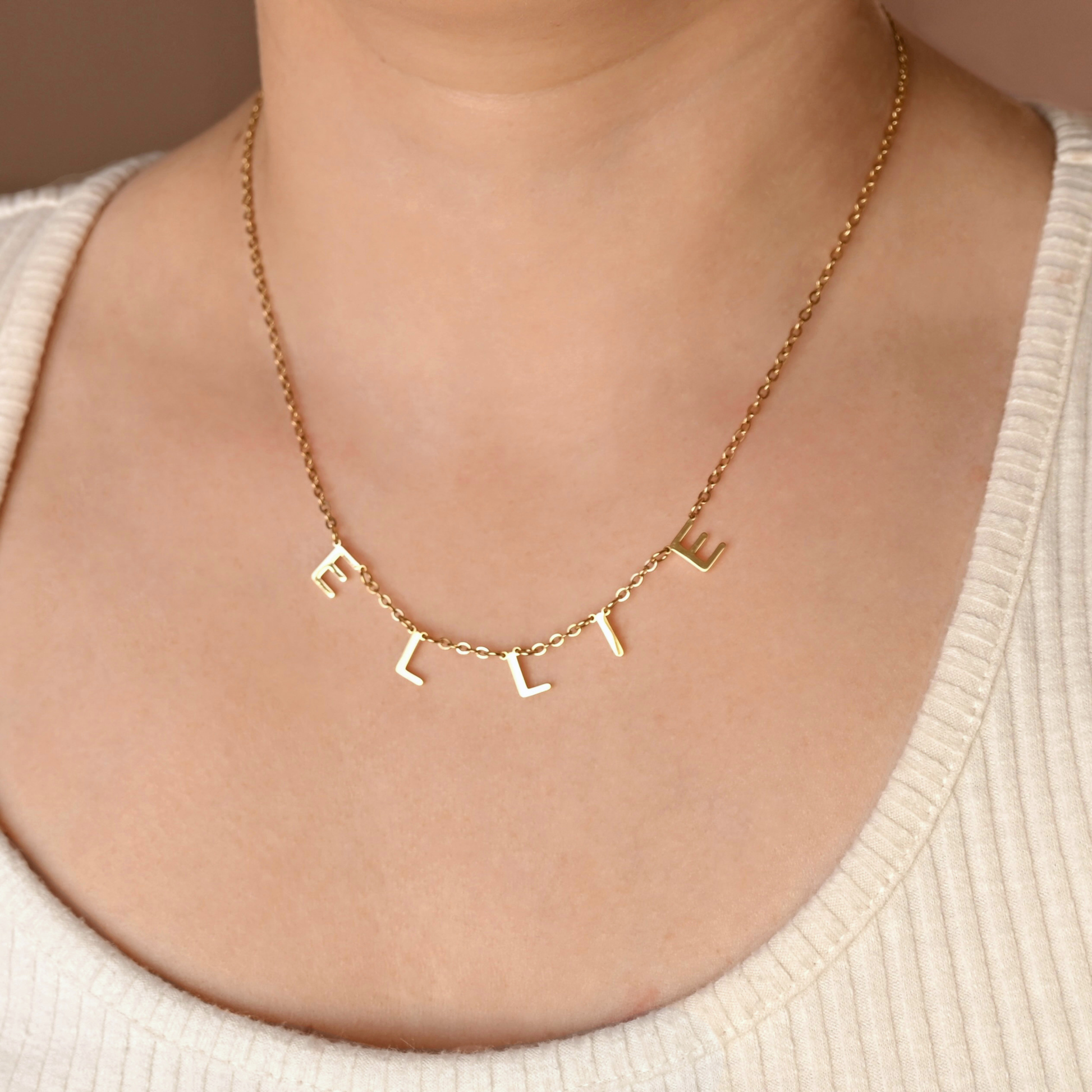 A close-up of a person's neck and chest wearing a delicate gold chain necklace with dangling letter charms spelling 'E L L I E'. The person is wearing a cream-colored ribbed top against a soft beige background.