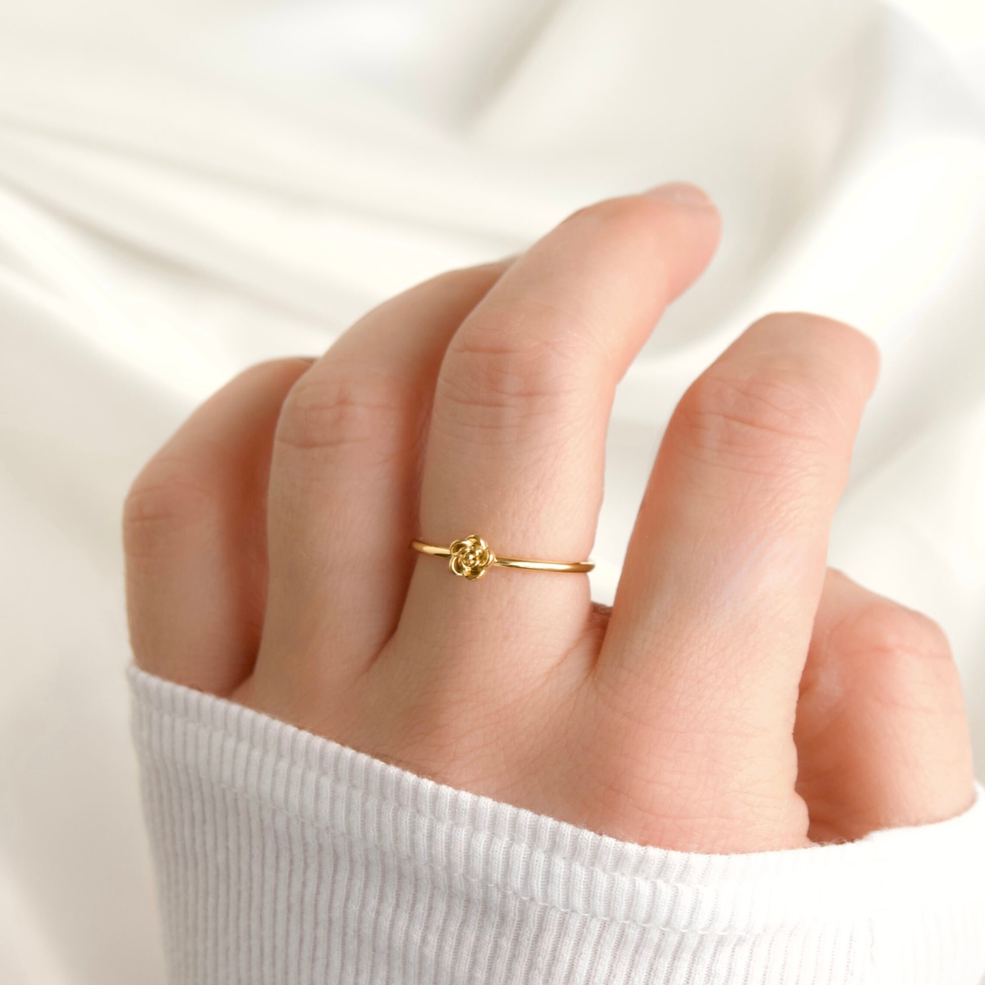 A delicate gold ring with a small floral or knot-like centerpiece, worn on a person's ring finger against a soft white fabric background.