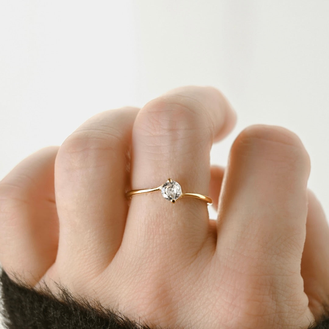 A delicate gold solitaire ring with a clear round gemstone worn on a person's ring finger, shown in a close-up shot against a soft, light background.