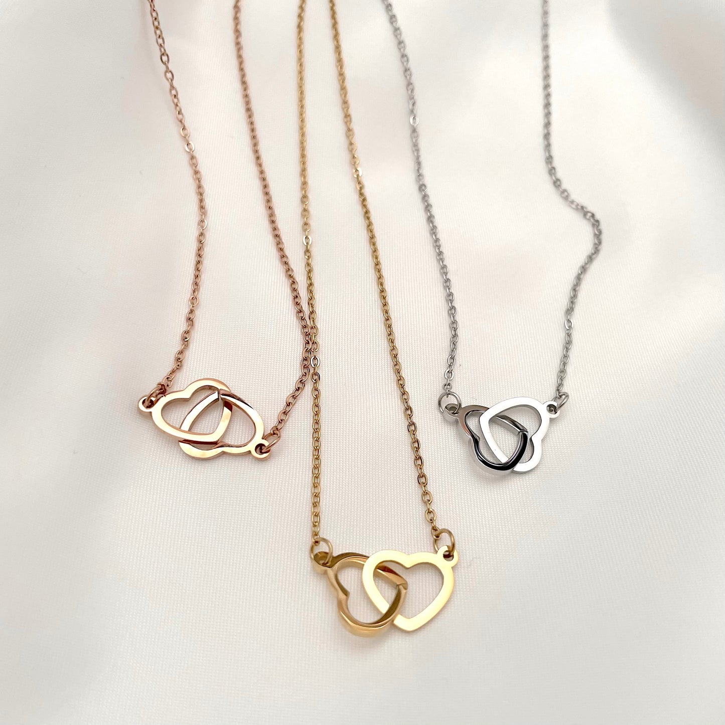 Top view of three interlocked heart necklaces in gold, rose gold and silver against a light background