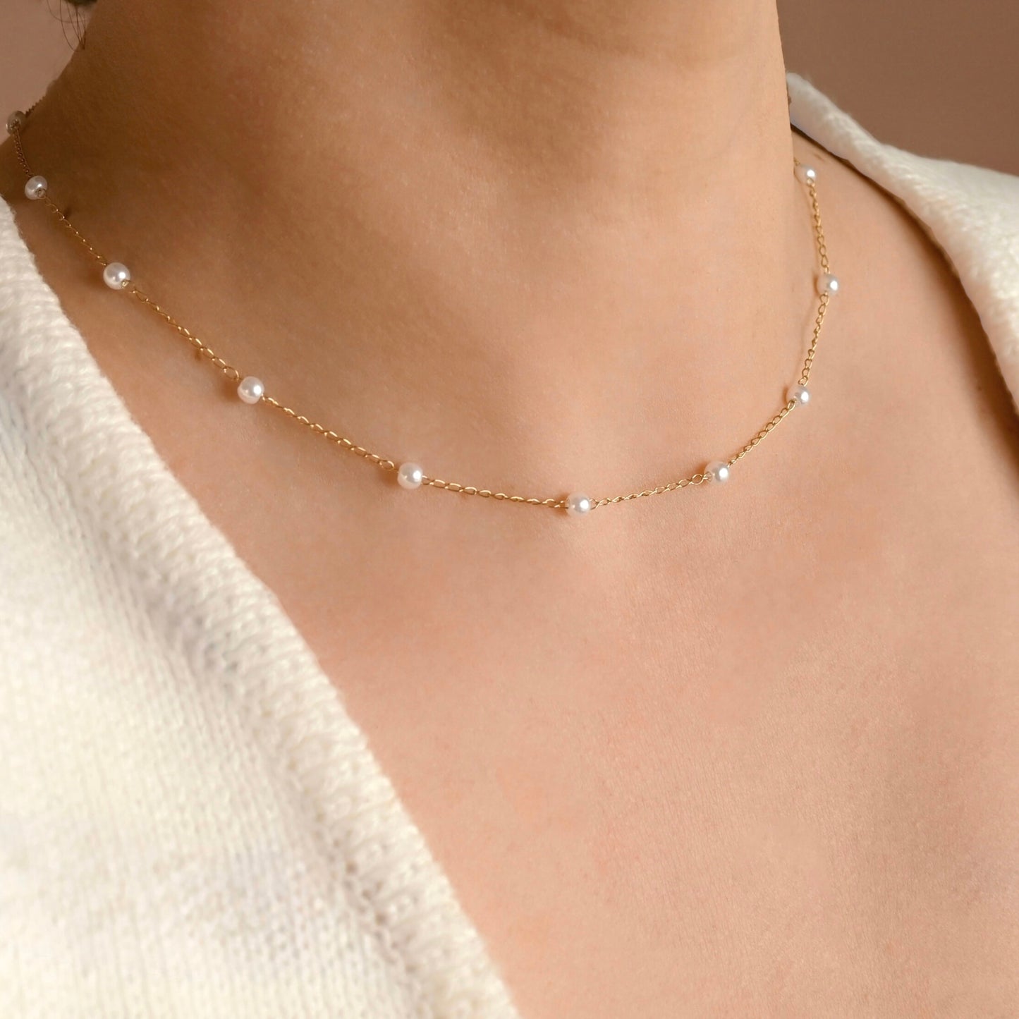 A delicate gold chain necklace with small white pearls spaced along the front, worn on a model's neck over a cream-colored knit top.