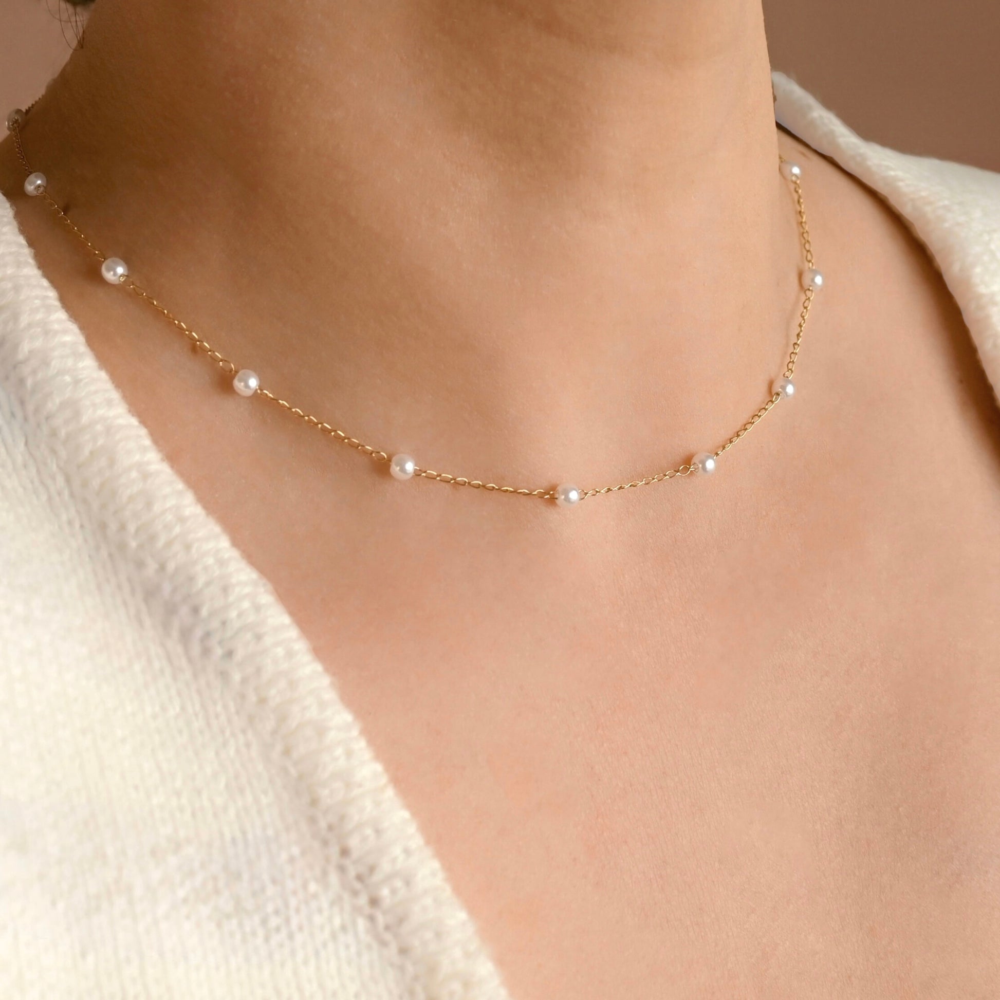 A delicate gold chain necklace with small white pearls spaced along the front, worn on a model's neck over a cream-colored knit top.