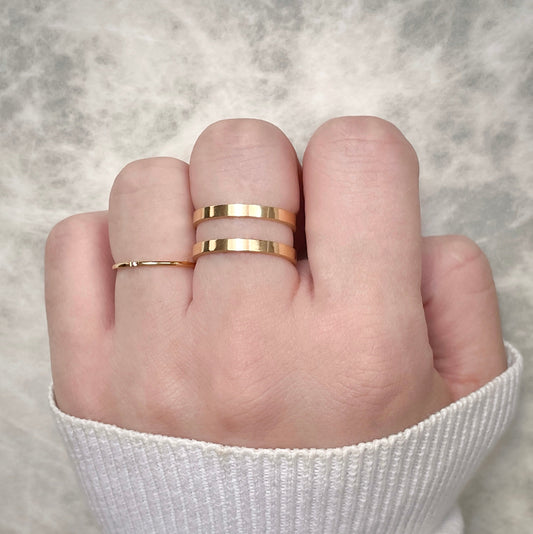 Close-up of a hand wearing two gold rings, one ultra slim with a tiny CZ, one bold and layered on a neutral background