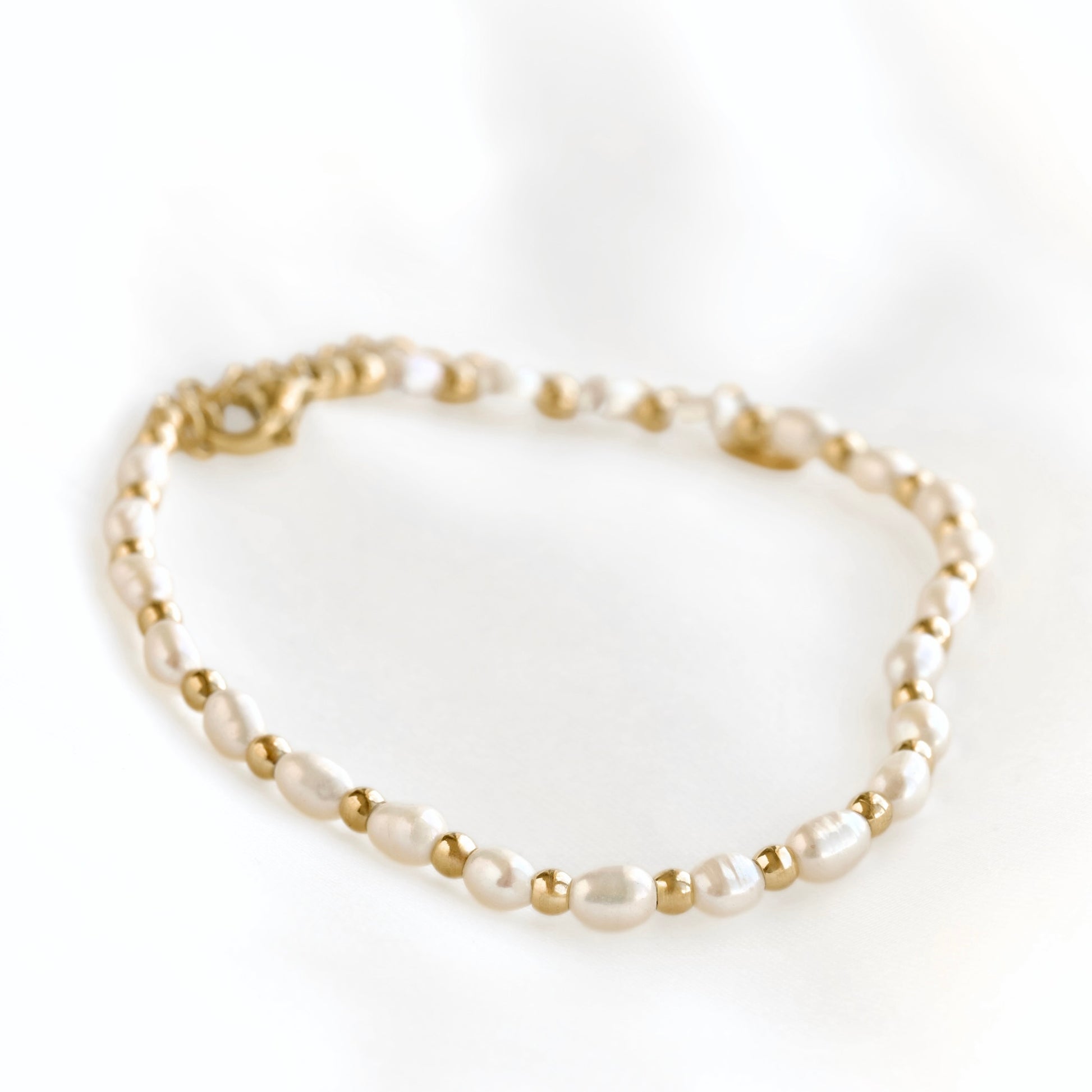 A delicate beaded bracelet featuring irregular white pearls separated by small gold spacer beads and a gold clasp, photographed on a soft white fabric background.