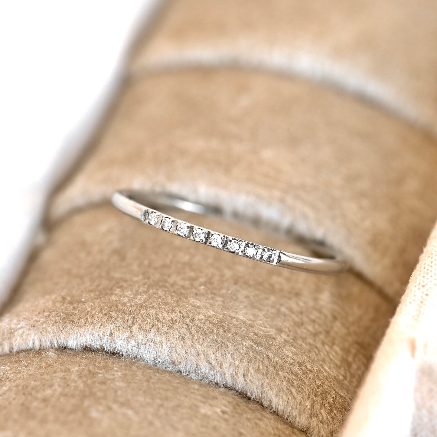 A thin silver ring with a row of small clear stones, displayed on a beige fabric ring holder.