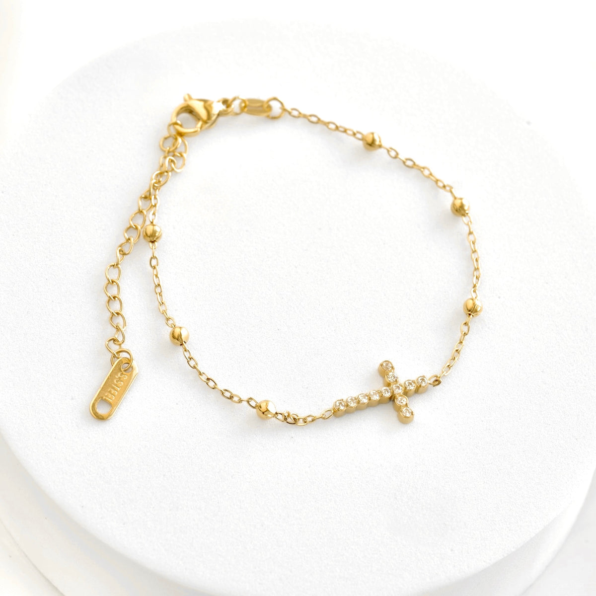 A delicate gold chain bracelet with a small cross charm set with tiny clear stones, displayed on a white circular jewelry stand.