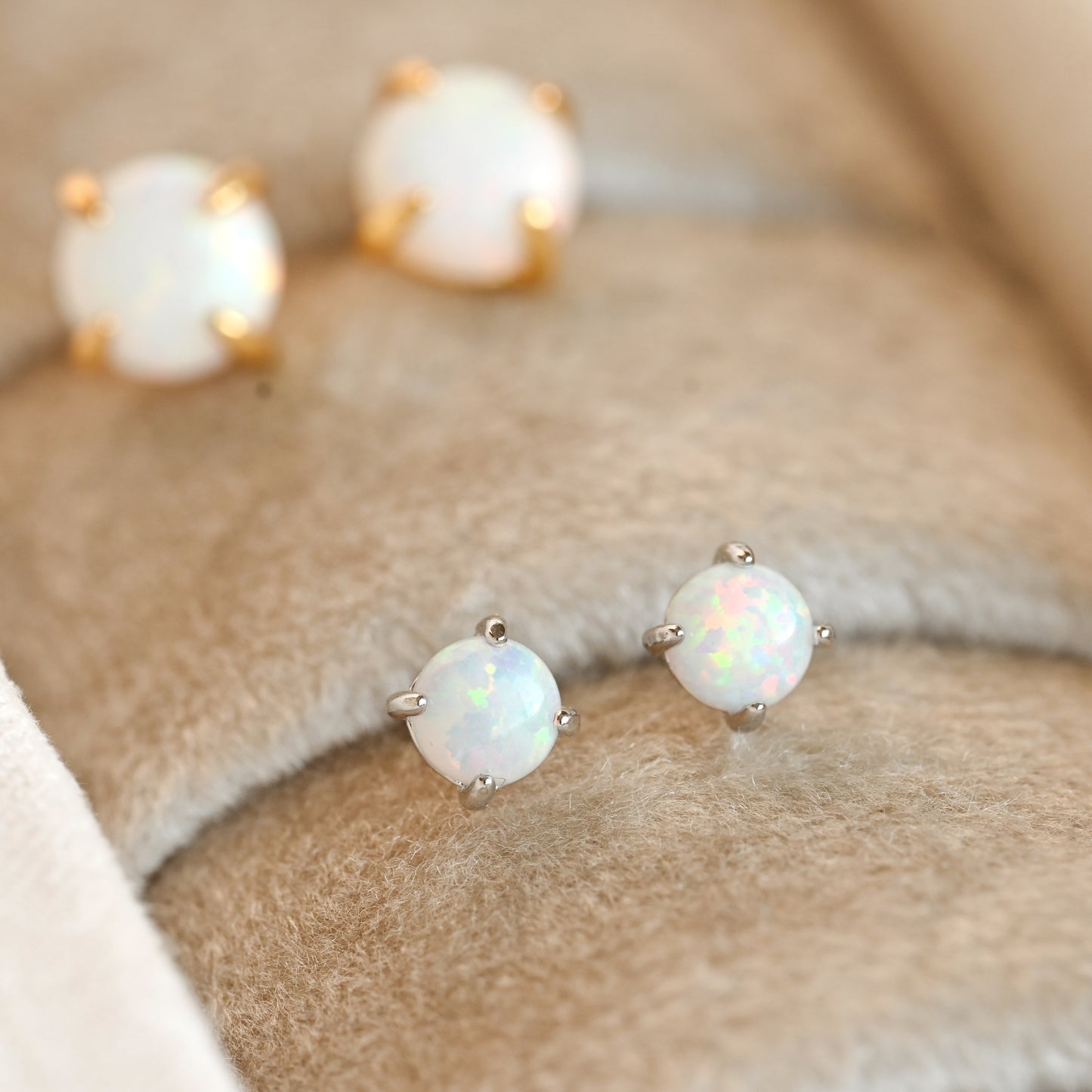 Two pairs of round white opal stud earrings with gold and silver prongs displayed on a soft beige velvet jewelry surface.