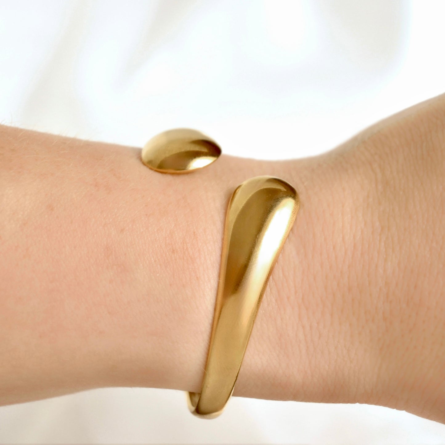 A polished gold cuff bracelet with a unique asymmetrical, curved design, shown worn on a person's wrist against a soft white background.