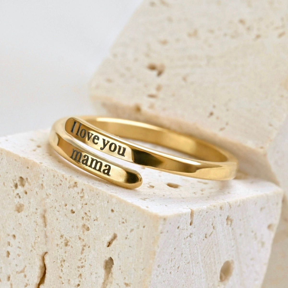 Dainty gold wrap ring on a textured beige stone block, with the top band engraved with the words 'I love you ' and bottom band with mama'.