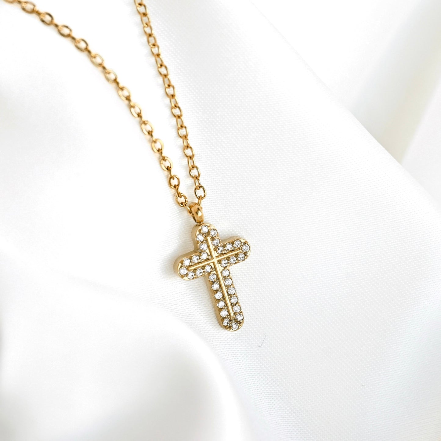 A gold cross pendant necklace with small clear rhinestones, resting on a draped white fabric background.