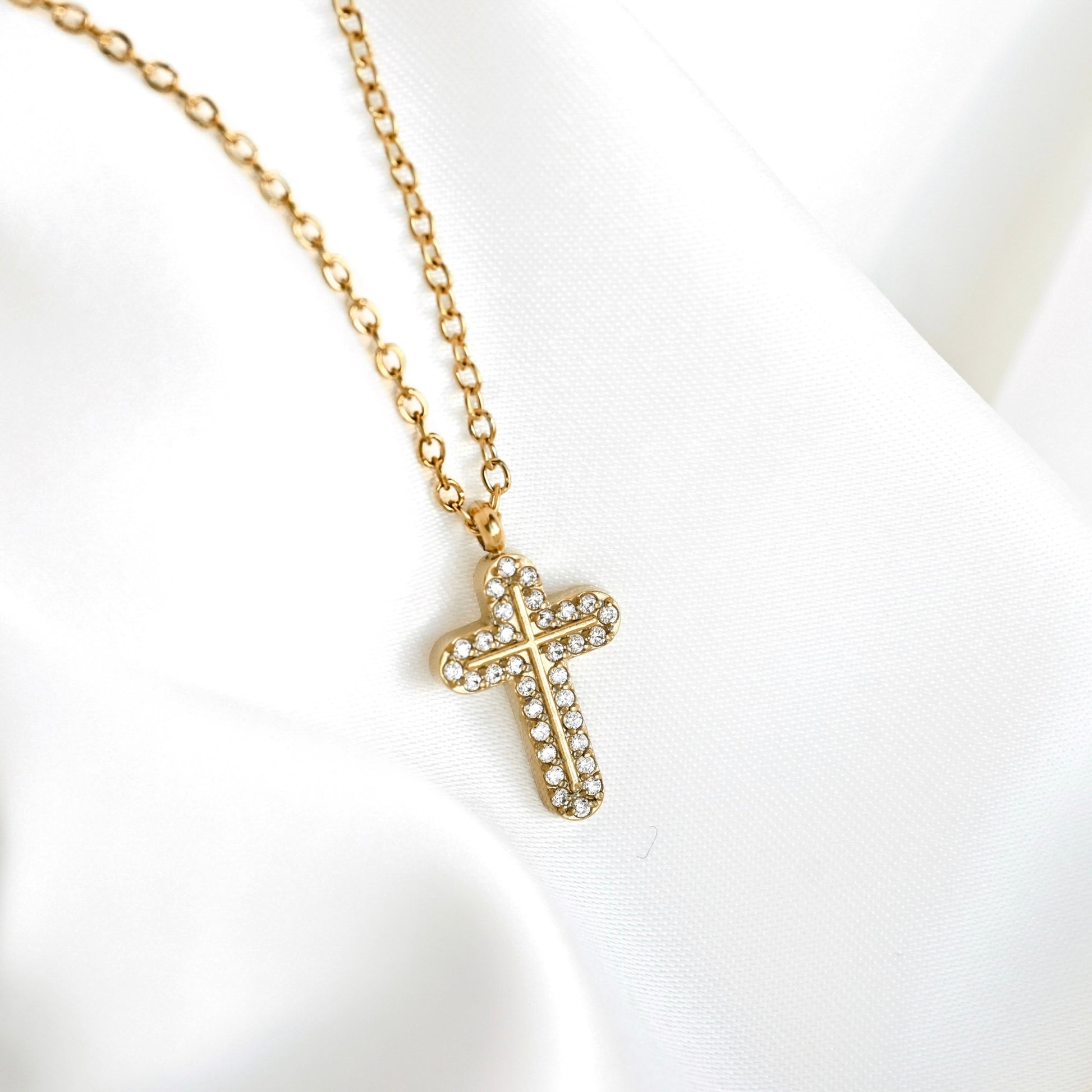 A gold cross pendant necklace with small clear rhinestones, resting on a draped white fabric background.