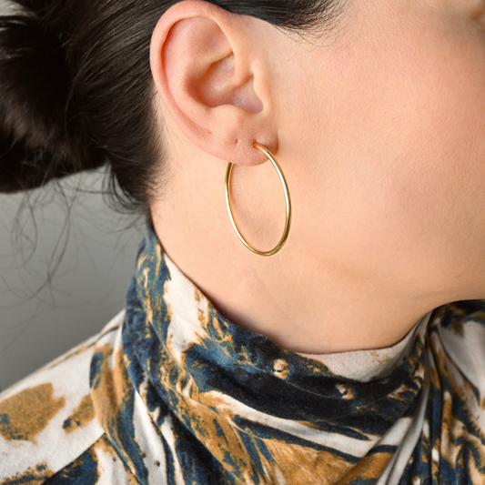 A close-up of a person's ear wearing medium sized thin, polished gold hoop earrings, paired with a patterned top in blue, gold, and white.