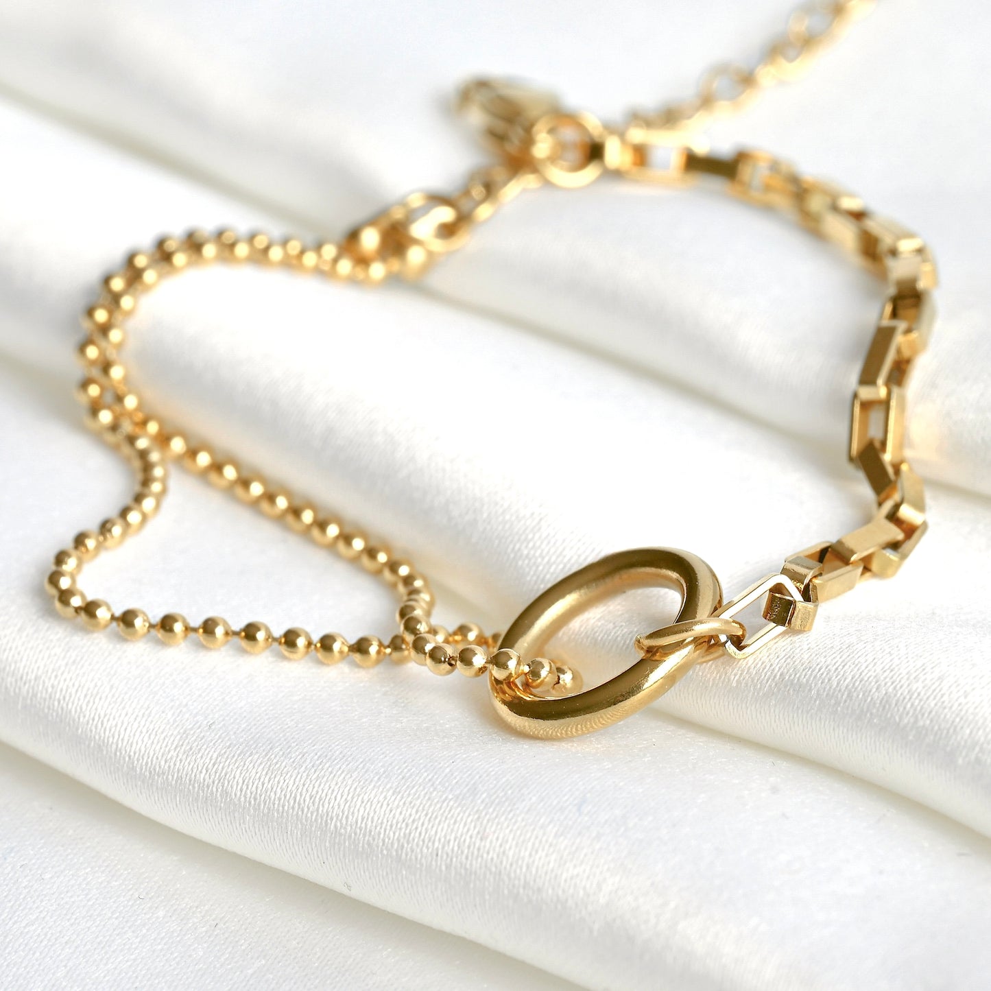 Close up view of gold chain bracelet with two different chains and a circle charm connector on a white fabric background