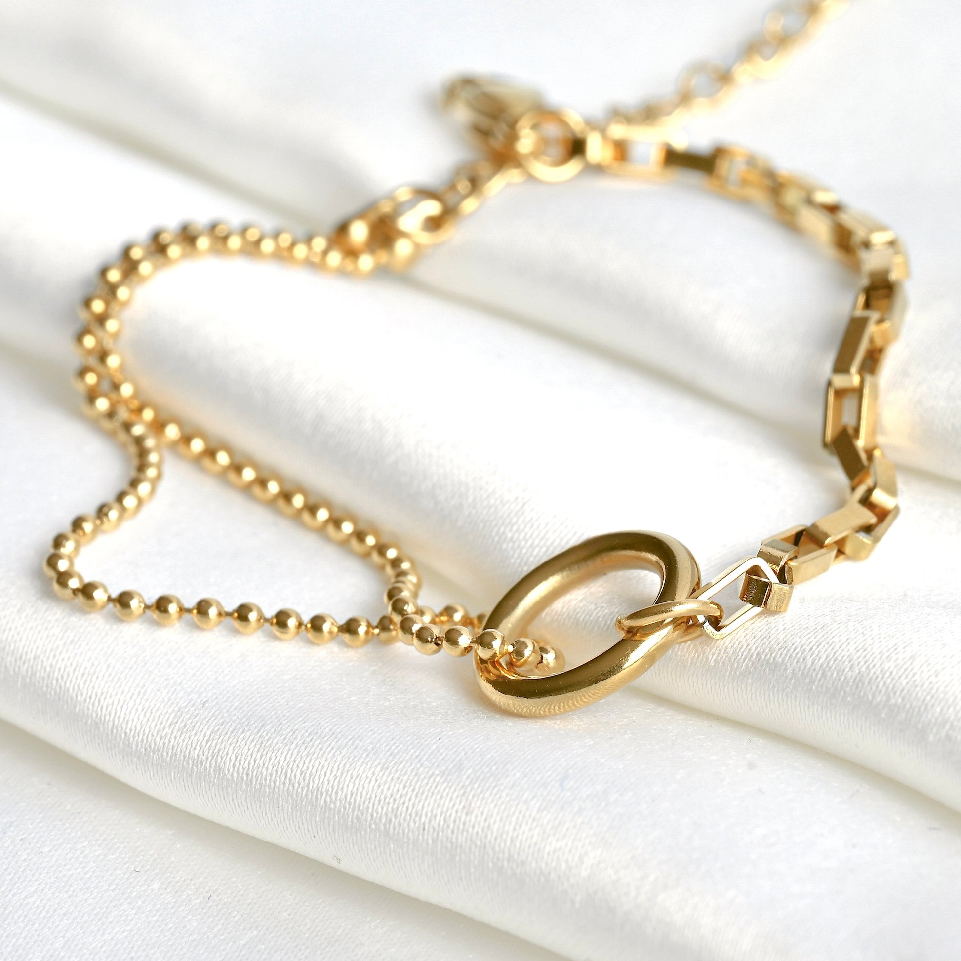 Close up view of gold chain bracelet with two different chains and a circle charm connector on a white fabric background