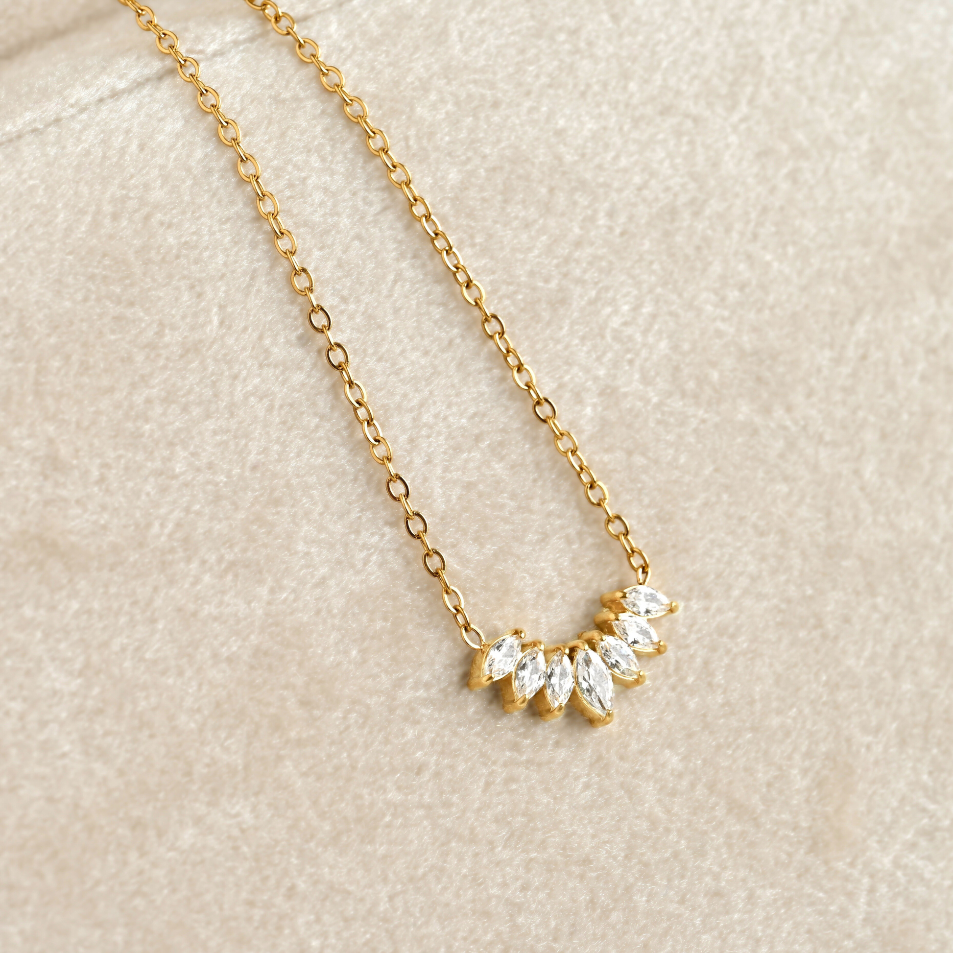 A delicate gold chain necklace featuring a small arched cluster of marquise-cut clear stones in a gold setting, displayed on a textured beige surface.