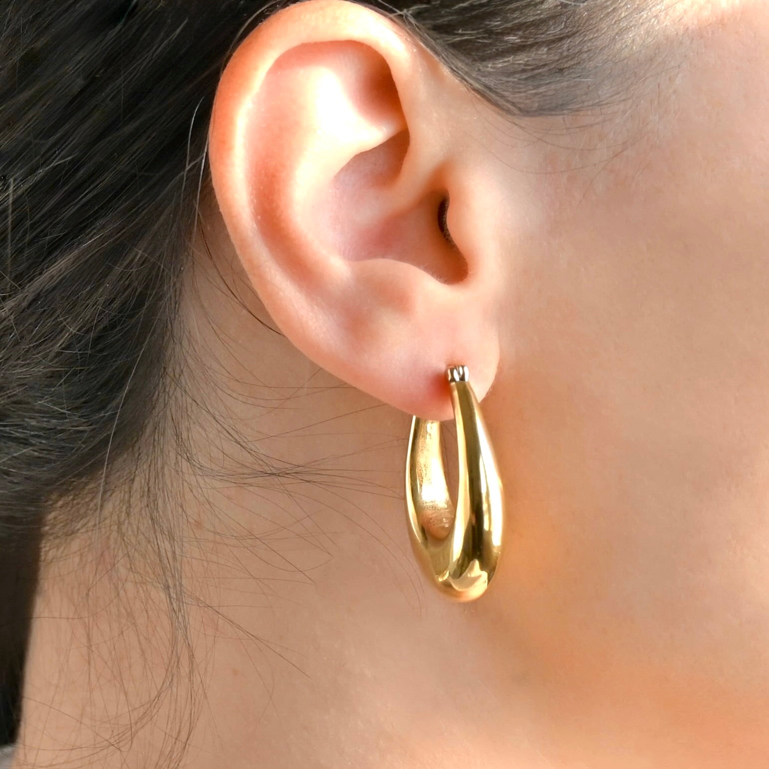 Thick gold drop hoop earrings worn by a person with dark hair.