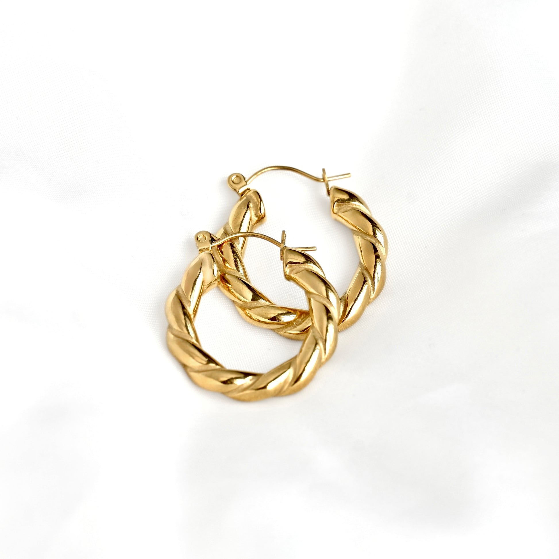 A pair of gold twisted hoop earrings with a polished finish, displayed on a soft white fabric background.