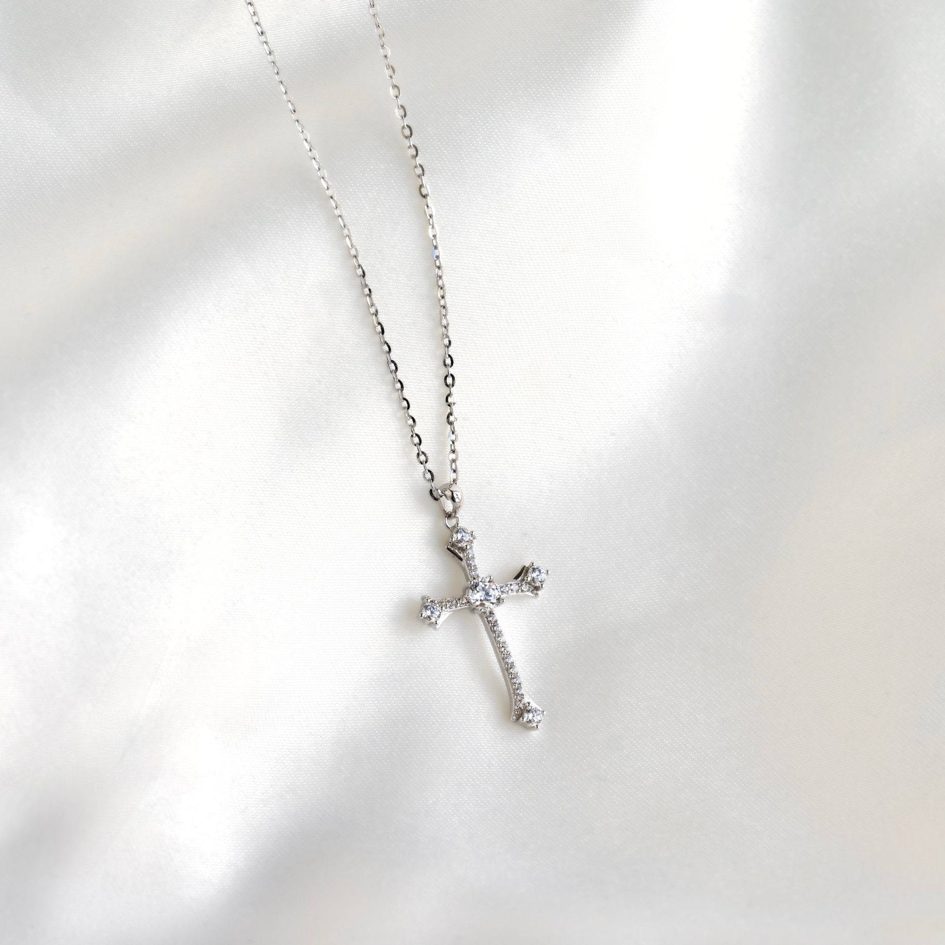 A silver cross pendant necklace adorned with small clear crystals, resting on a soft, white satin fabric background.