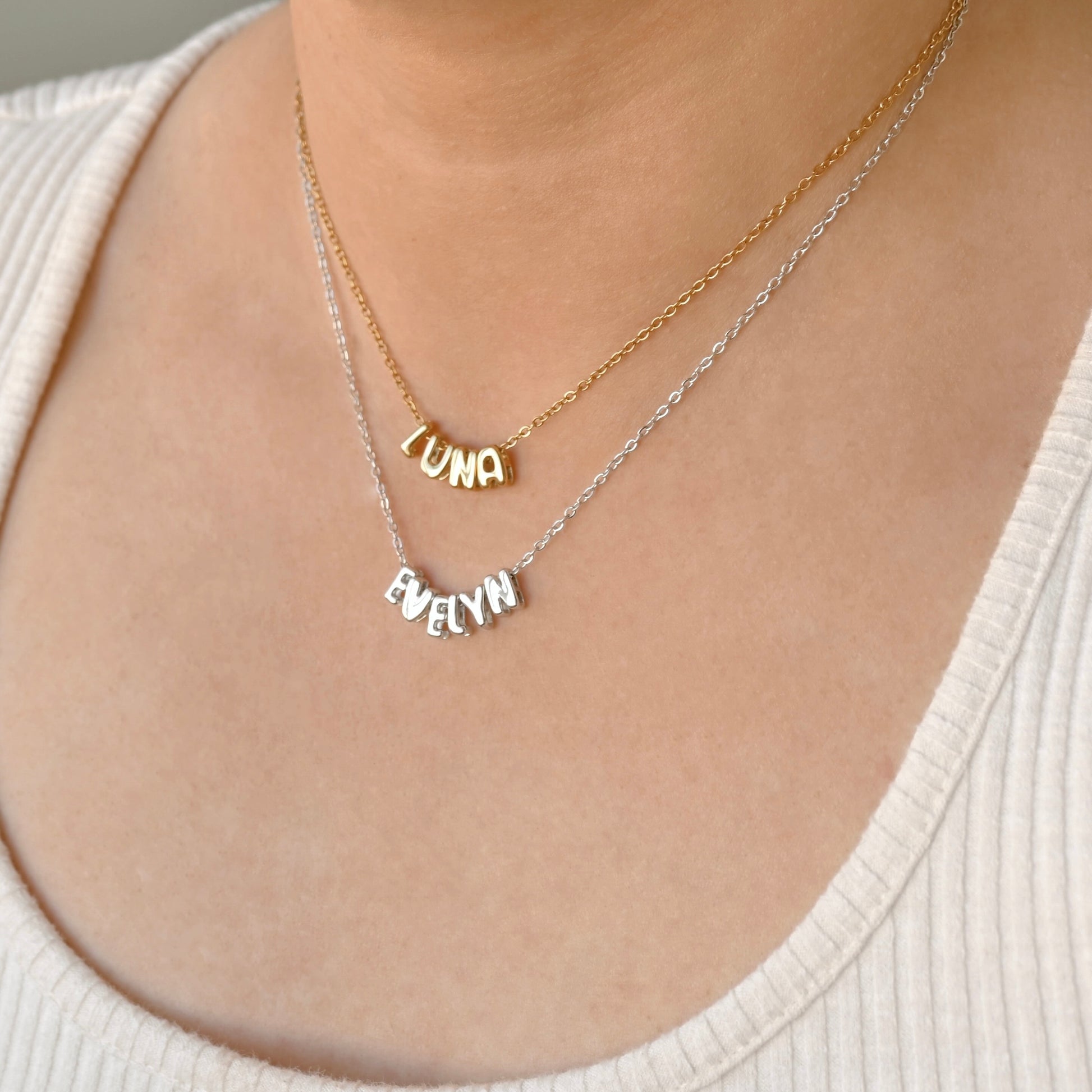 A close-up of a person wearing two delicate necklaces: a gold chain with a 'LUNA' nameplate and a silver chain with a 'EVELYN' nameplate, both displayed on the chest over a cream ribbed top.