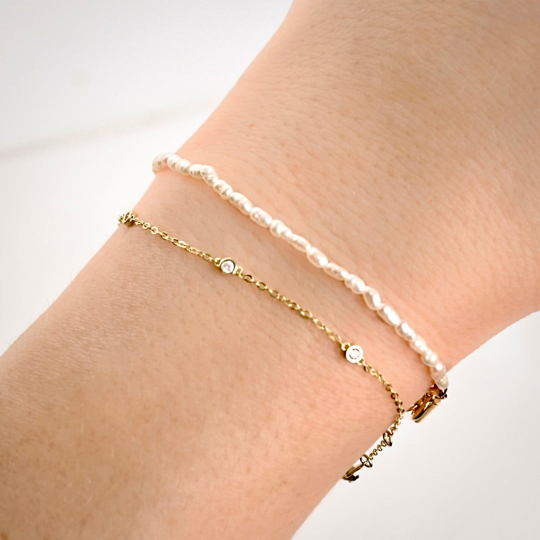 Two delicate bracelets worn on a wrist: a thin gold chain bracelet with small clear stone accents and a white pearl beaded bracelet, shown against a soft light background.