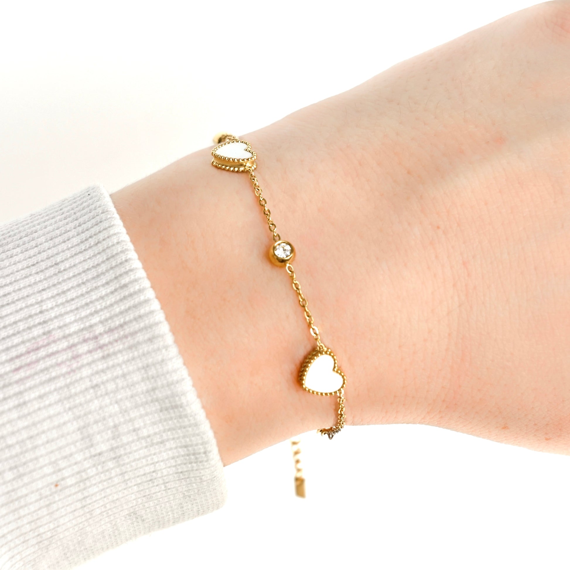 A delicate gold chain bracelet with two heart charms featuring a white mother of pearl inlay with a clear bezel stone in between, shown worn on a person's wrist