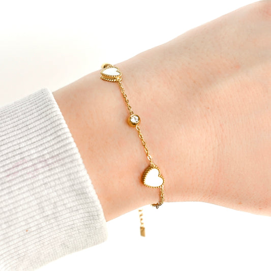 A delicate gold chain bracelet with two heart charms featuring a white mother of pearl inlay with a clear bezel stone in between, shown worn on a person's wrist
