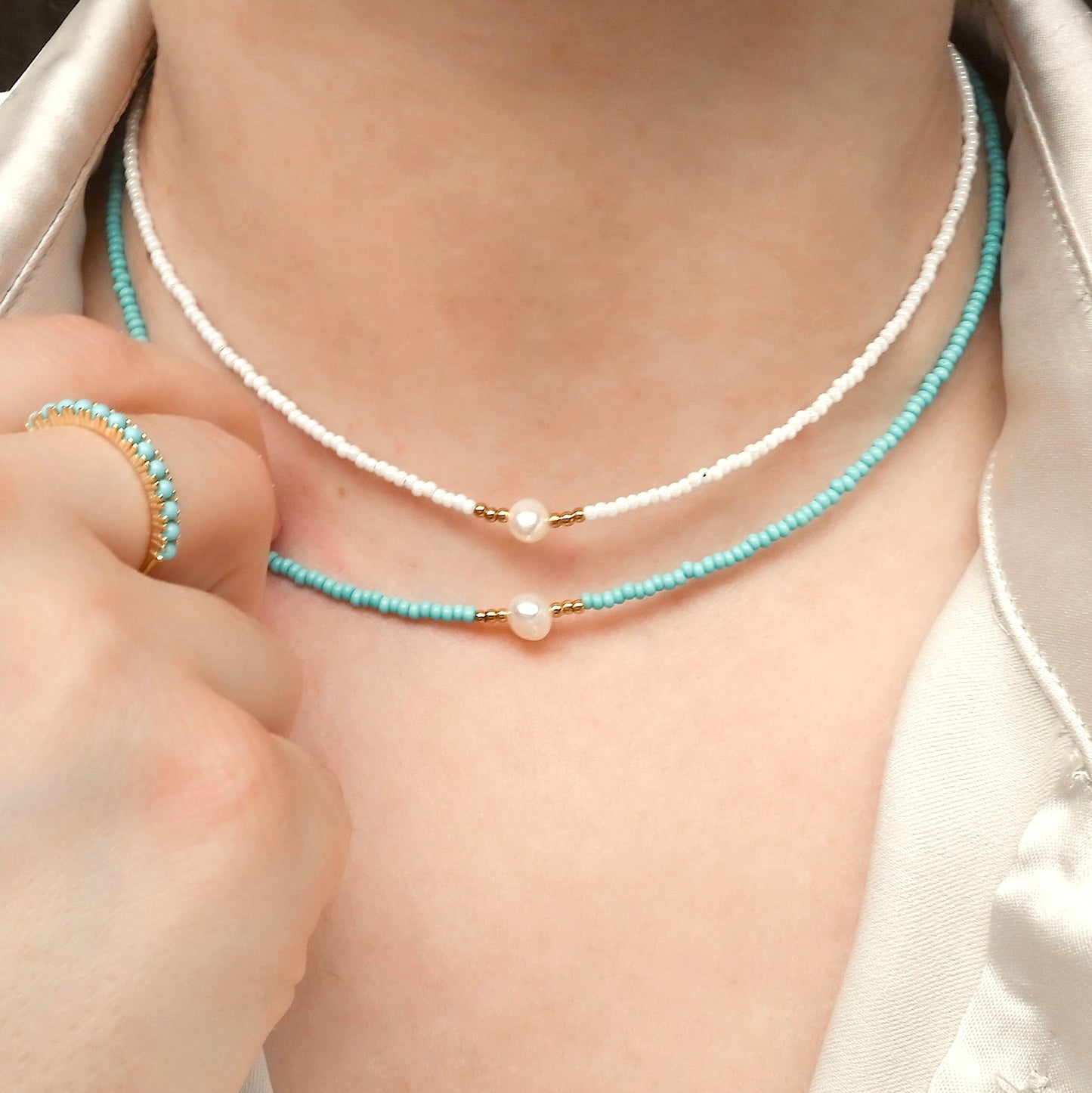 Close-up of a person wearing two freshwater pearl necklaces, one with white and one with turquoise seed beads, with a turquoise gemstone ring on a neutral background