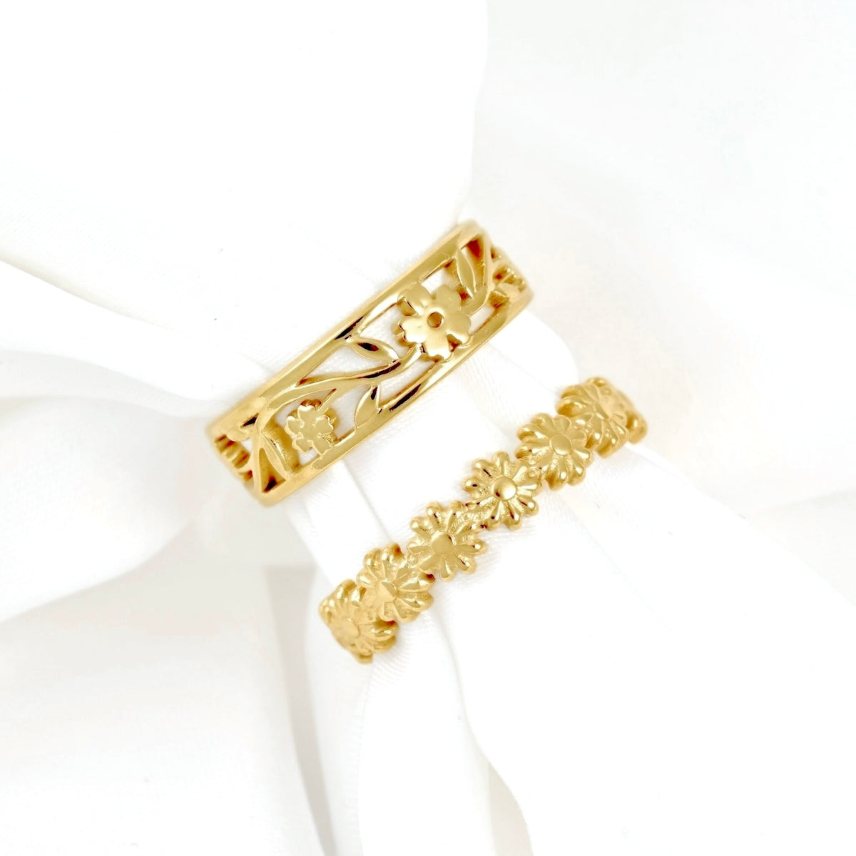 Two gold rings displayed on a white fabric ring holder. The top ring is a wide band with an openwork floral and leaf pattern, while the bottom ring is a delicate, continuous band featuring a repeating floral motif.
