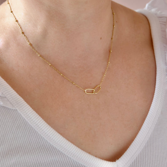 A delicate gold chain necklace with a small open-link paperclip-style pendant, shown worn on a model's neck over a white ribbed top.