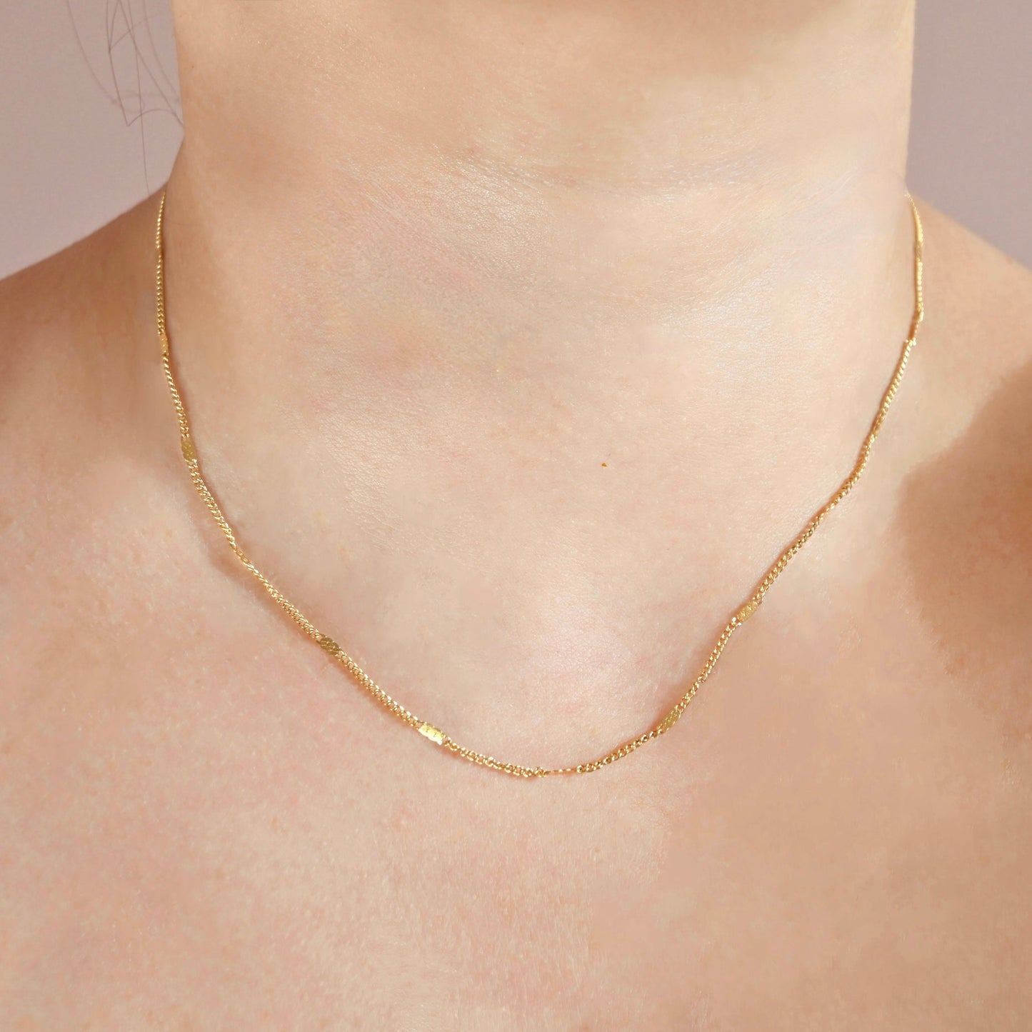 A delicate gold chain necklace worn on a person's neck, shown in a close-up shot against a soft, light pink background.