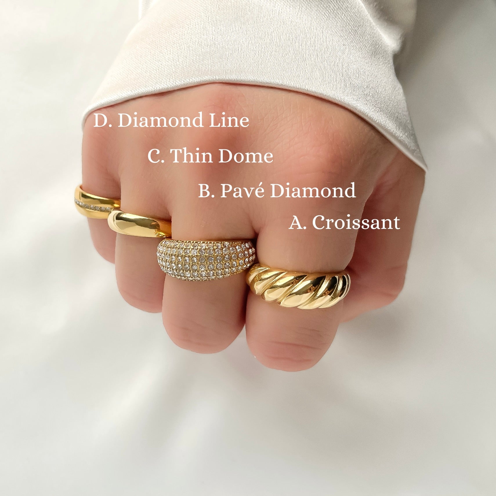 Hand wearing four gold domerings with different designs including diamond line, thin dome, pave diamond, and croissant designs with their text labels on a light background