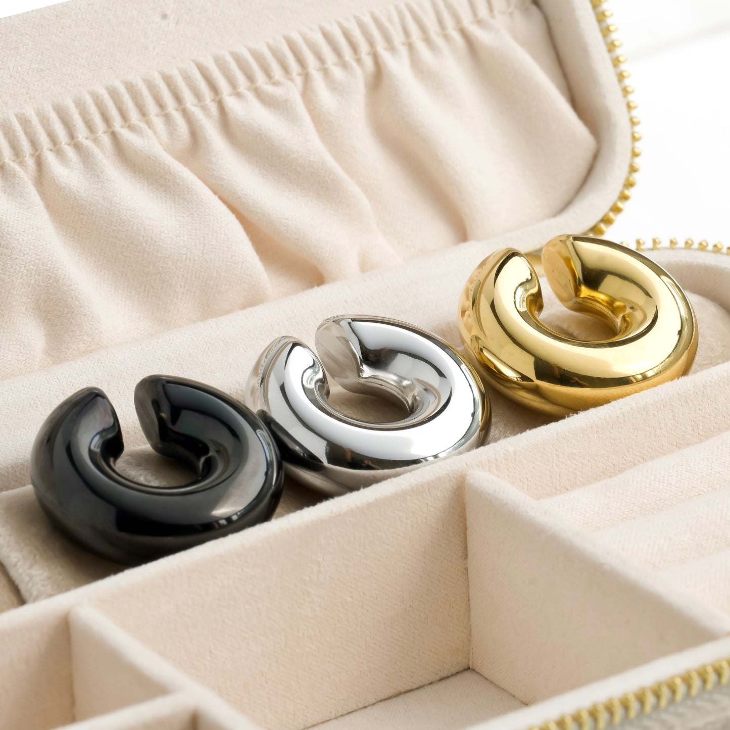 Three chunky cuff earrings in black, silver, and gold in a jewelry box.