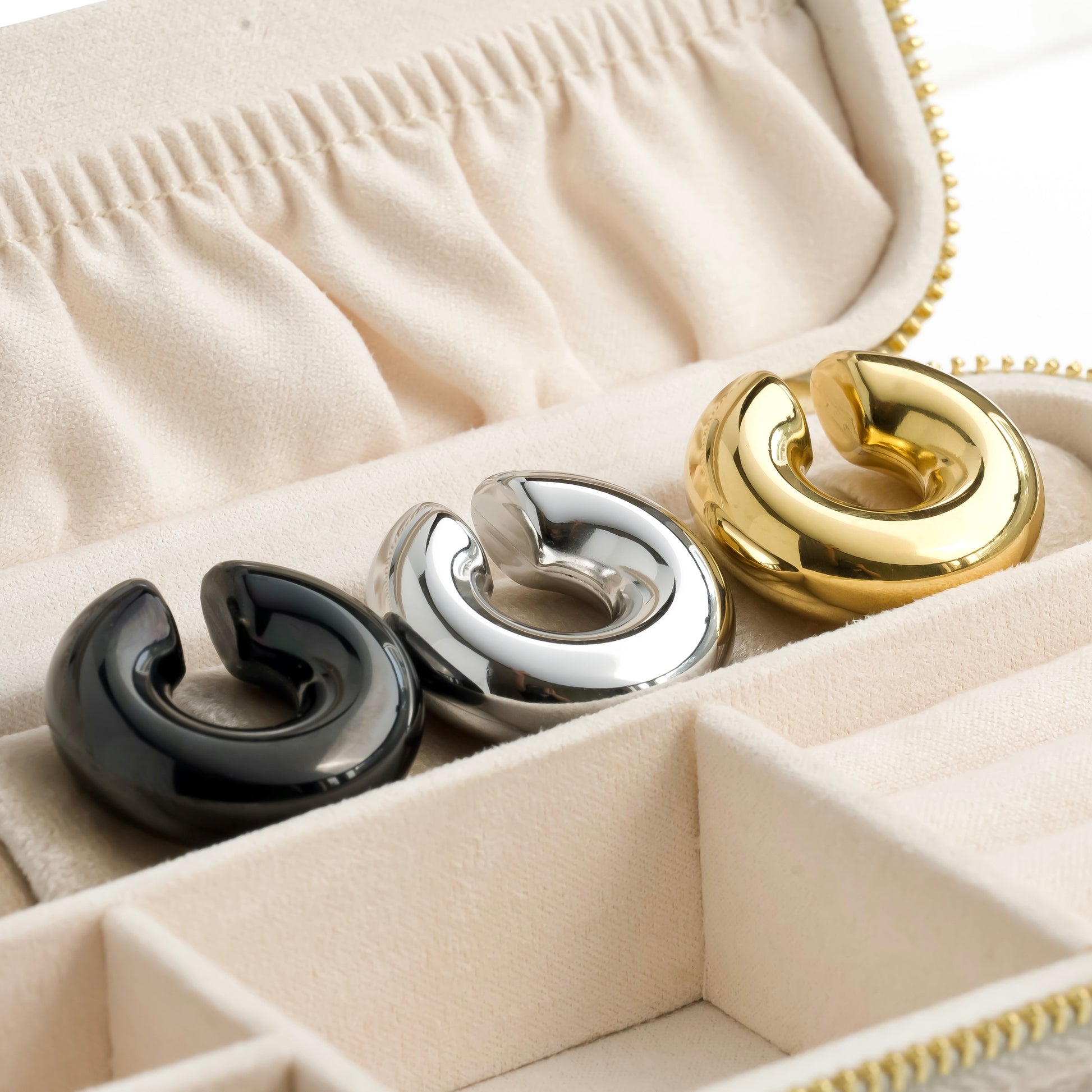 Three chunky cuff earrings in black, silver, and gold in a jewelry box.